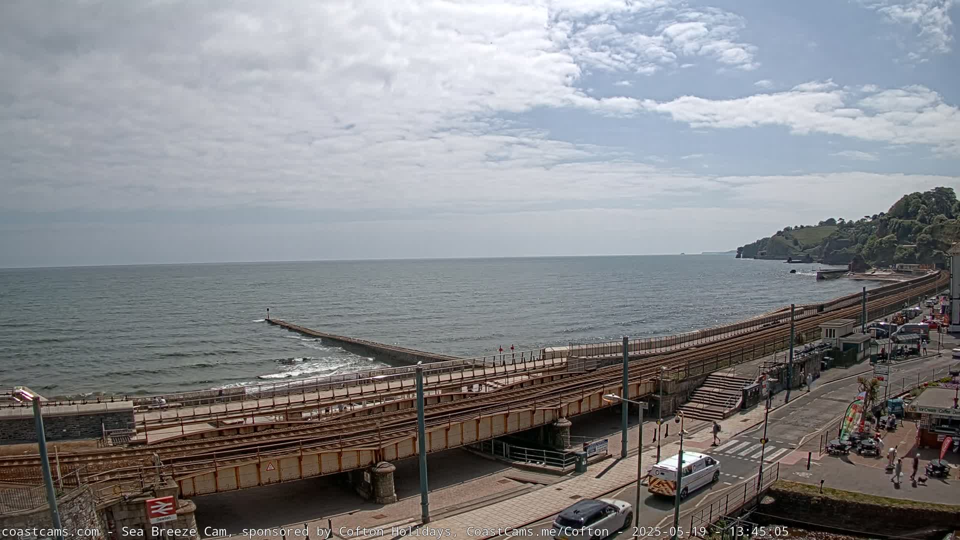 A mostly cloudy day shows a coastal scene with a railway line and seawall next to a roadway, all overlooking a somewhat choppy sea.