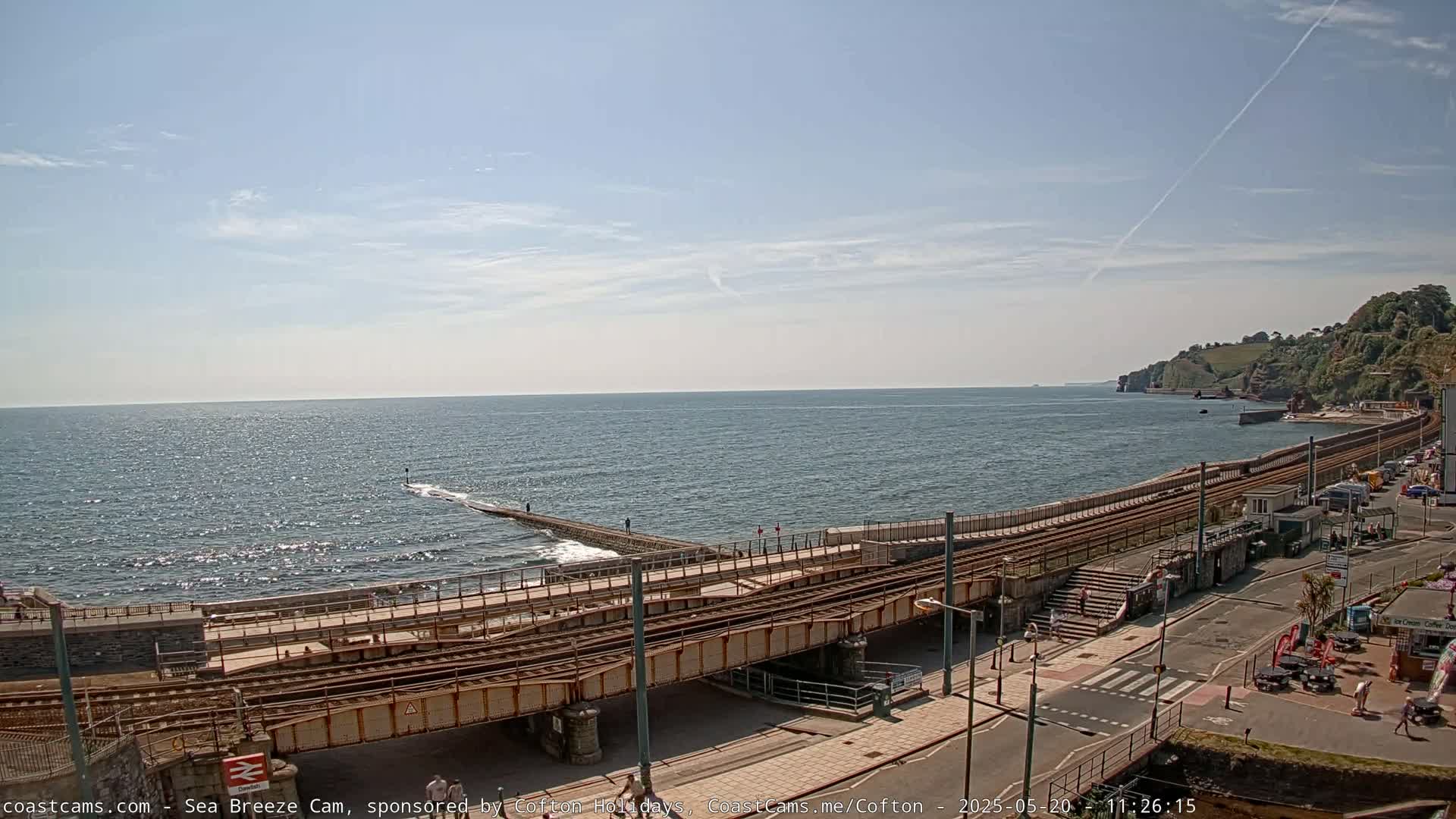A coastal scene on a sunny day shows a railway line crossing a seawall and leading to a town, with a calm sea and distant cliffs visible.