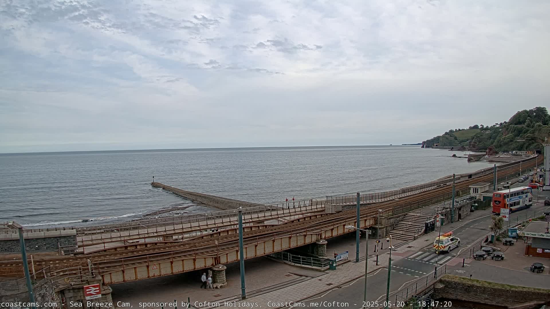 A coastal scene under a cloudy sky shows a railway track crossing a paved area next to a beach and calm ocean, with a small pier extending into the water and a bus parked on a nearby street.