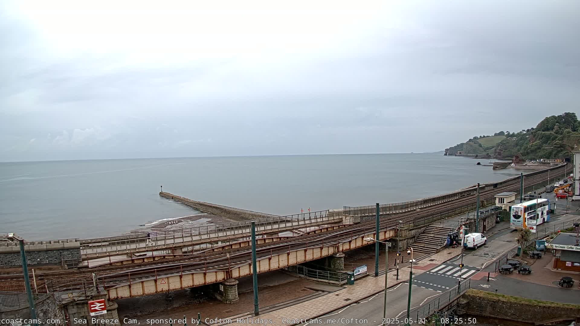 A coastal railway line and a breakwater extend into a calm sea under an overcast sky.