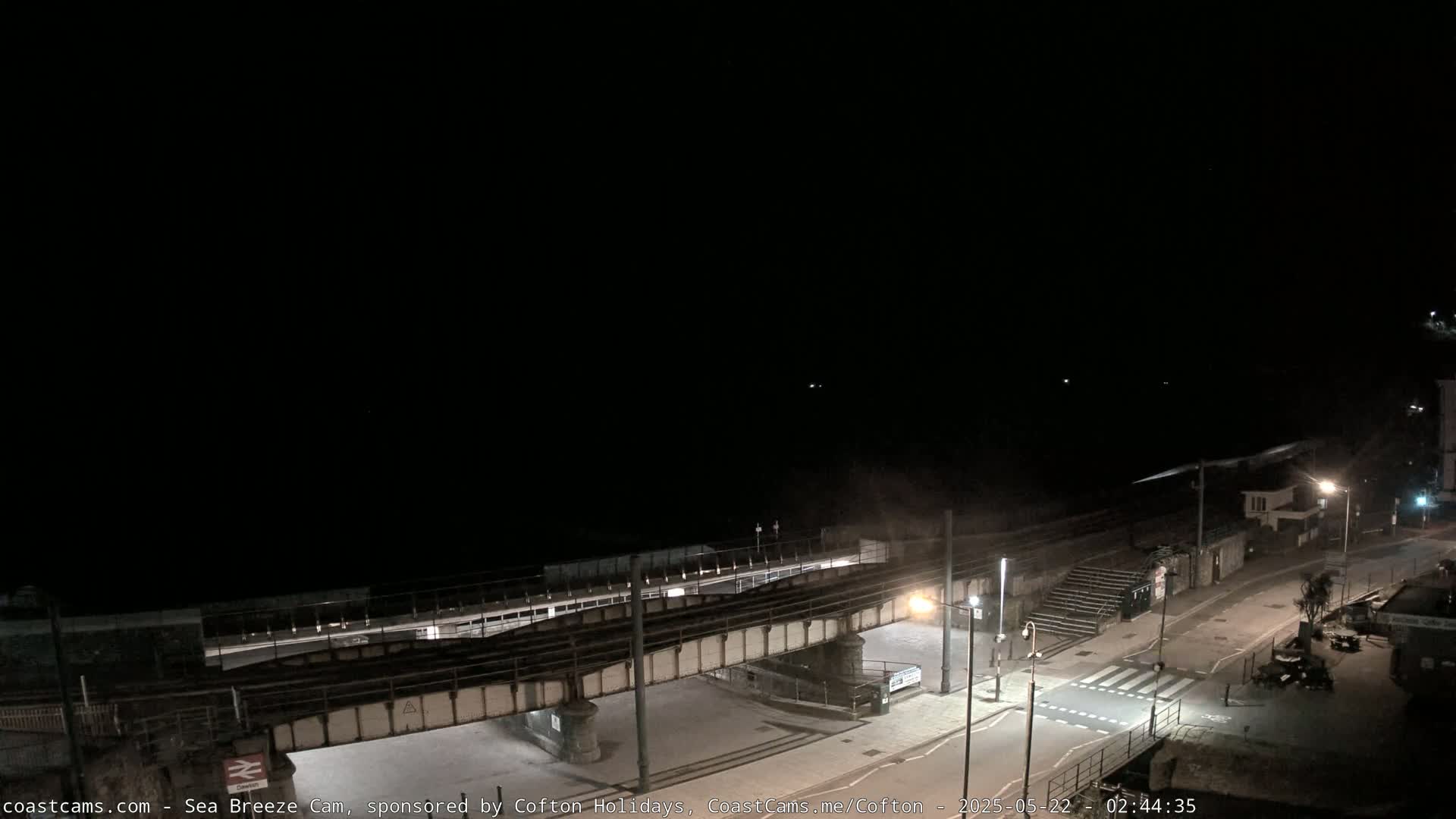A nighttime, coastal scene shows a train trestle over a road leading to a beach, under a dark sky.