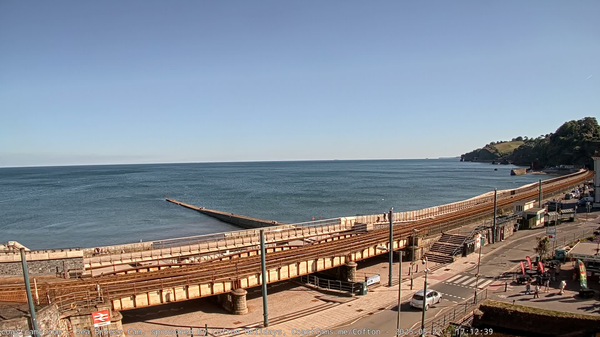 A seaside town scene on a sunny day features a railway line running along a seawall, with a calm ocean extending to the horizon.