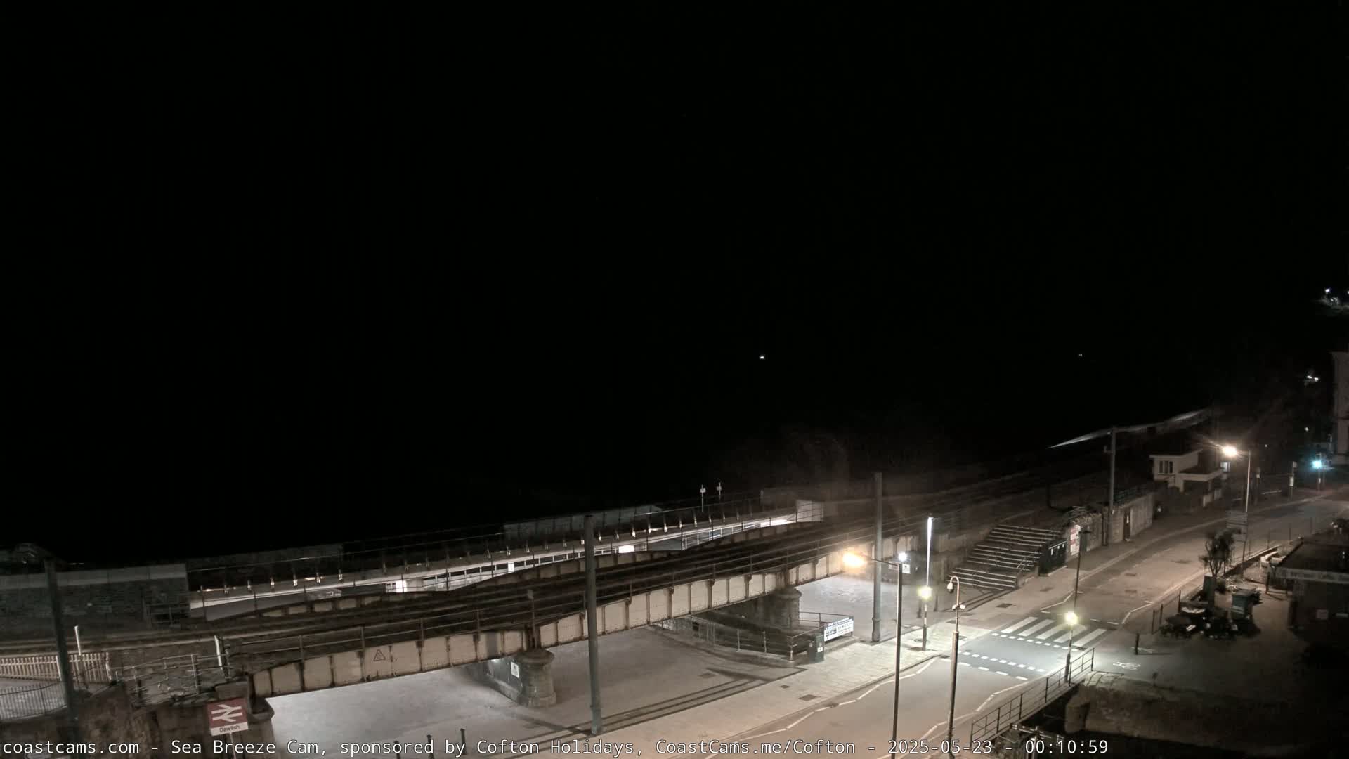 A nighttime view of a coastal town shows a train trestle over a road leading to a beach under a dark sky.