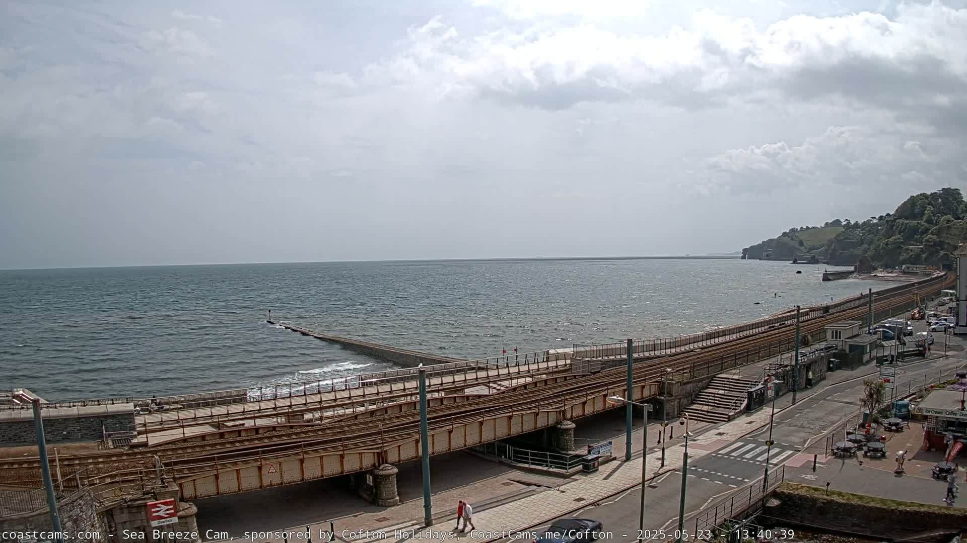 A coastal scene under a mostly cloudy sky shows a railway line running alongside a sea wall and a road, with a calm sea in the background.
