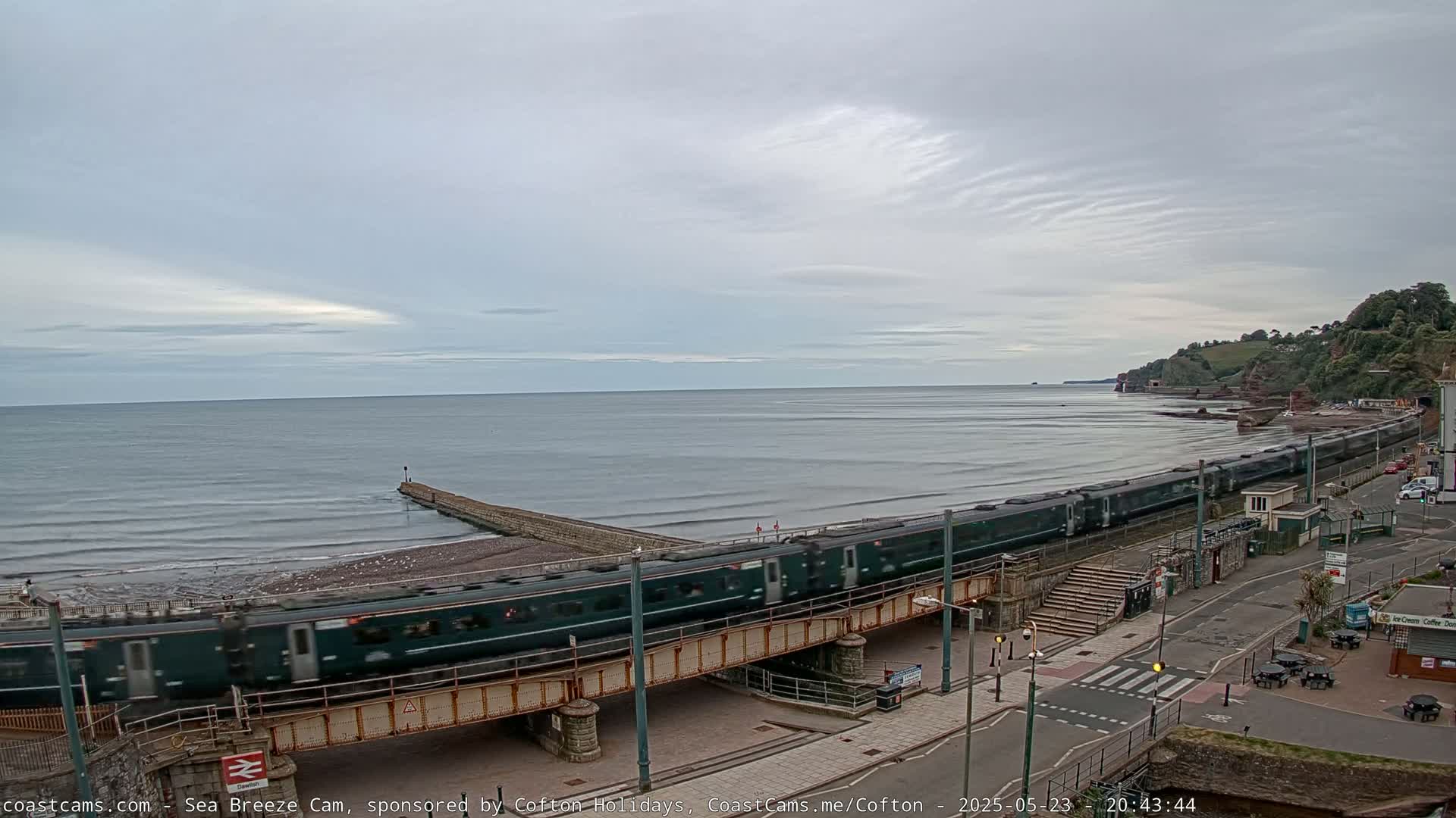 A long green train crosses a bridge over a beach beside a calm sea under a cloudy sky.