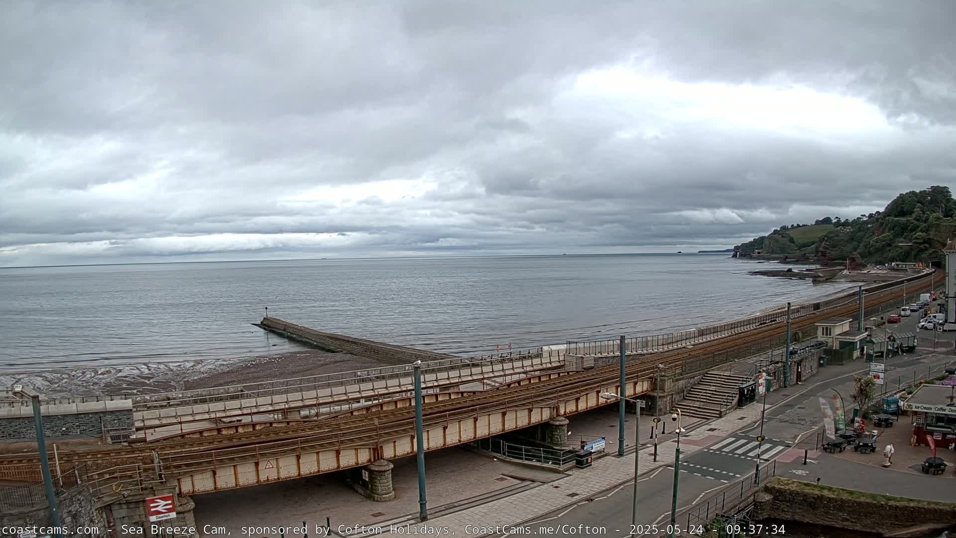 A railway track crosses over a beach and seaside road under an overcast sky.