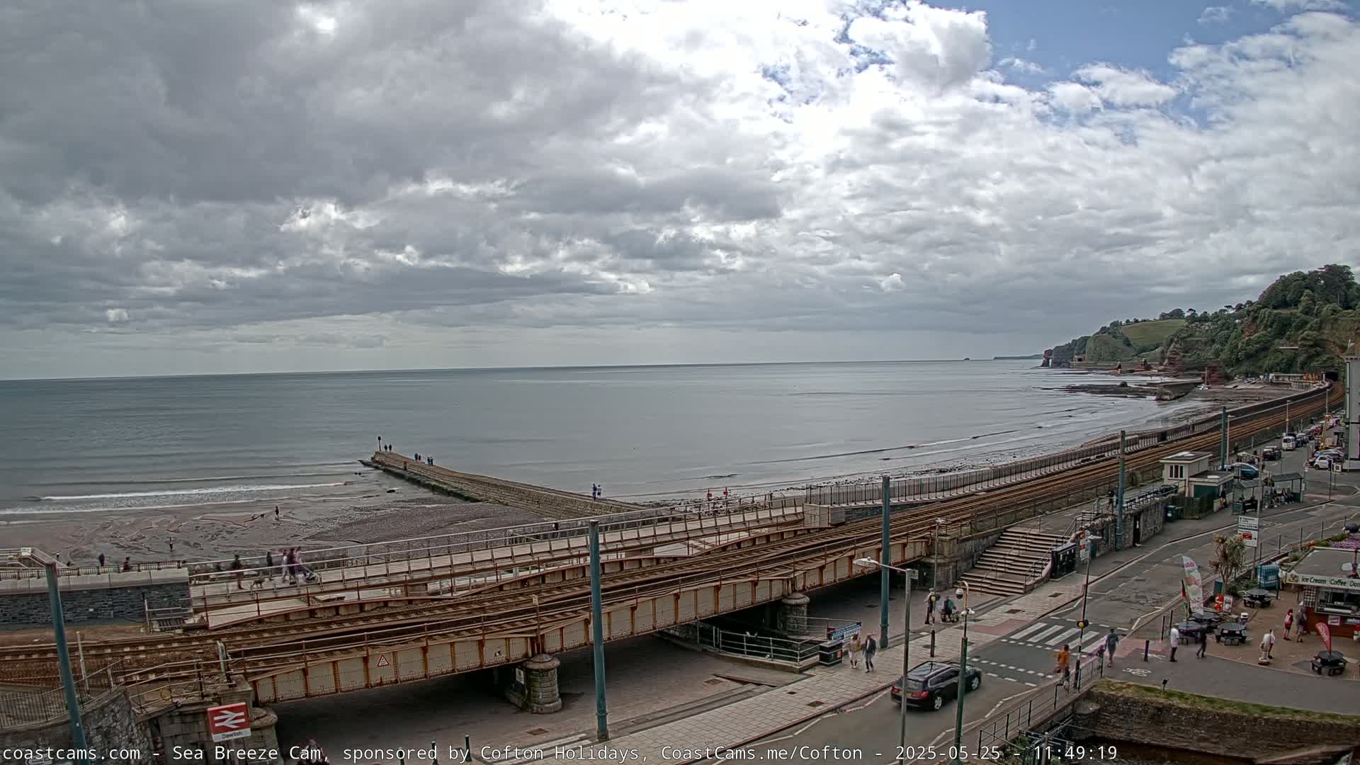An overcast day at a seaside town shows a railway track crossing a beach and a paved road with people walking along the shore and street.