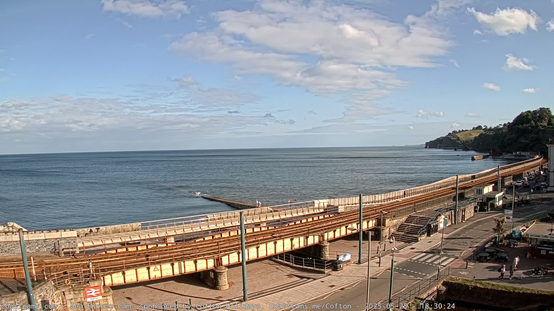 A coastal railway track runs along a seawall overlooking a calm ocean under a partly cloudy sky.