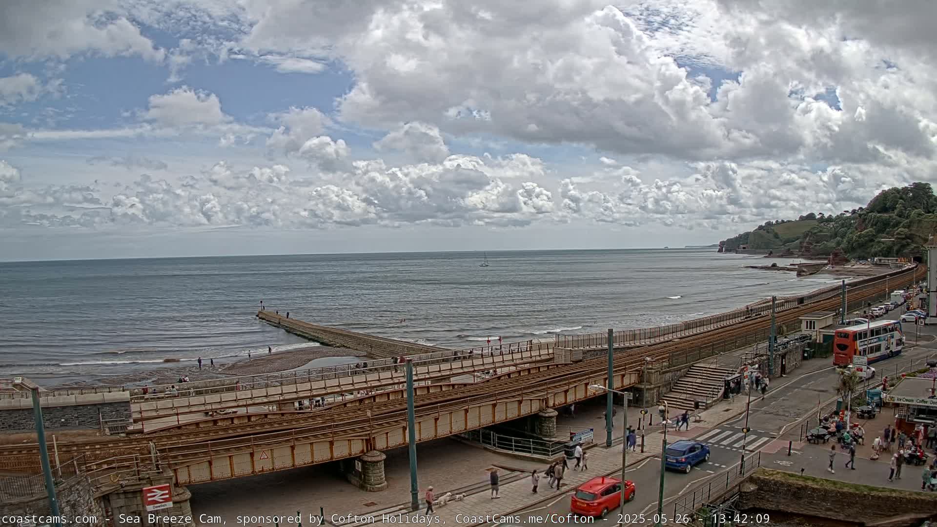 A coastal scene under a partly cloudy sky shows a railway line traversing a beach with people walking along the shore and a red car on the road next to it.