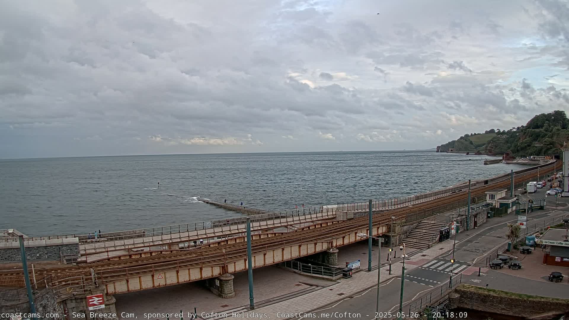 A coastal scene under a cloudy sky shows a railway track crossing a sea wall next to a paved road with buildings and cars.