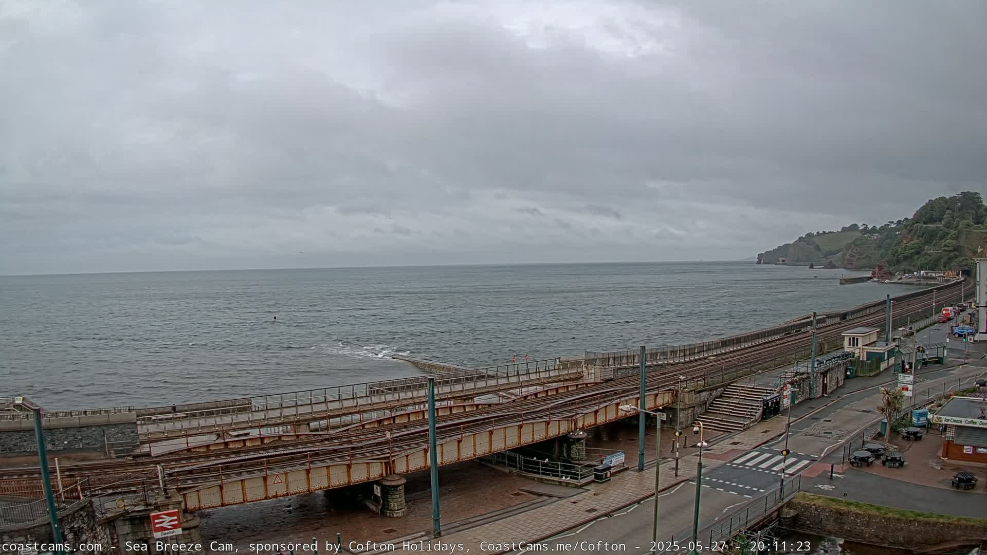 A choppy sea stretches to a cloudy sky, overlooking a coastal railway line and a road with buildings and some outdoor seating.