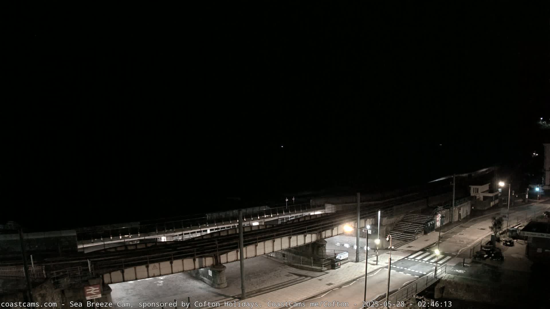A nighttime view of a coastal town, showing a railway bridge over a road and a dark ocean in the background under a clear sky.