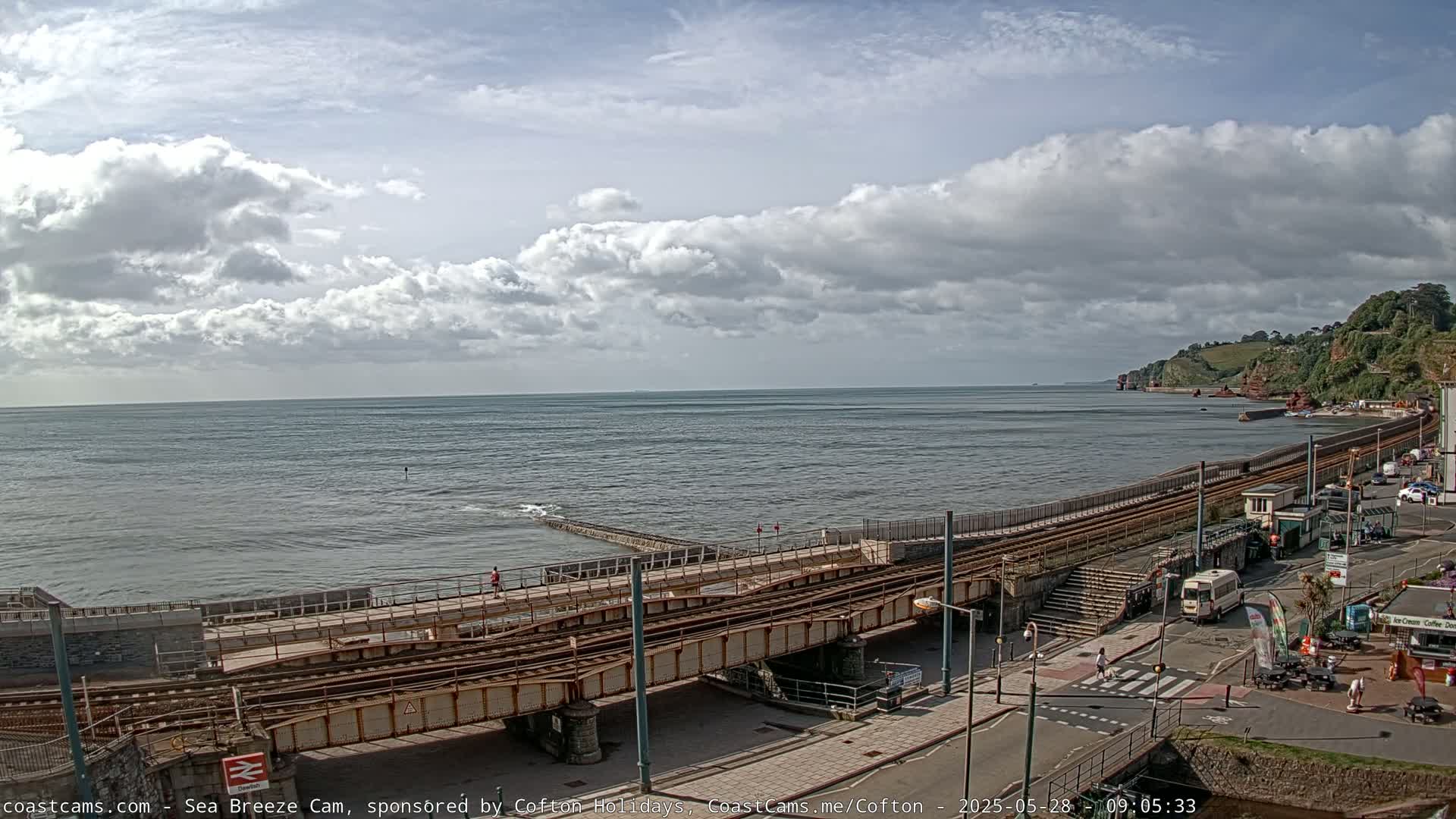 A partly cloudy day shows a coastal scene with train tracks running alongside a sea wall and a road, overlooking a calm sea.