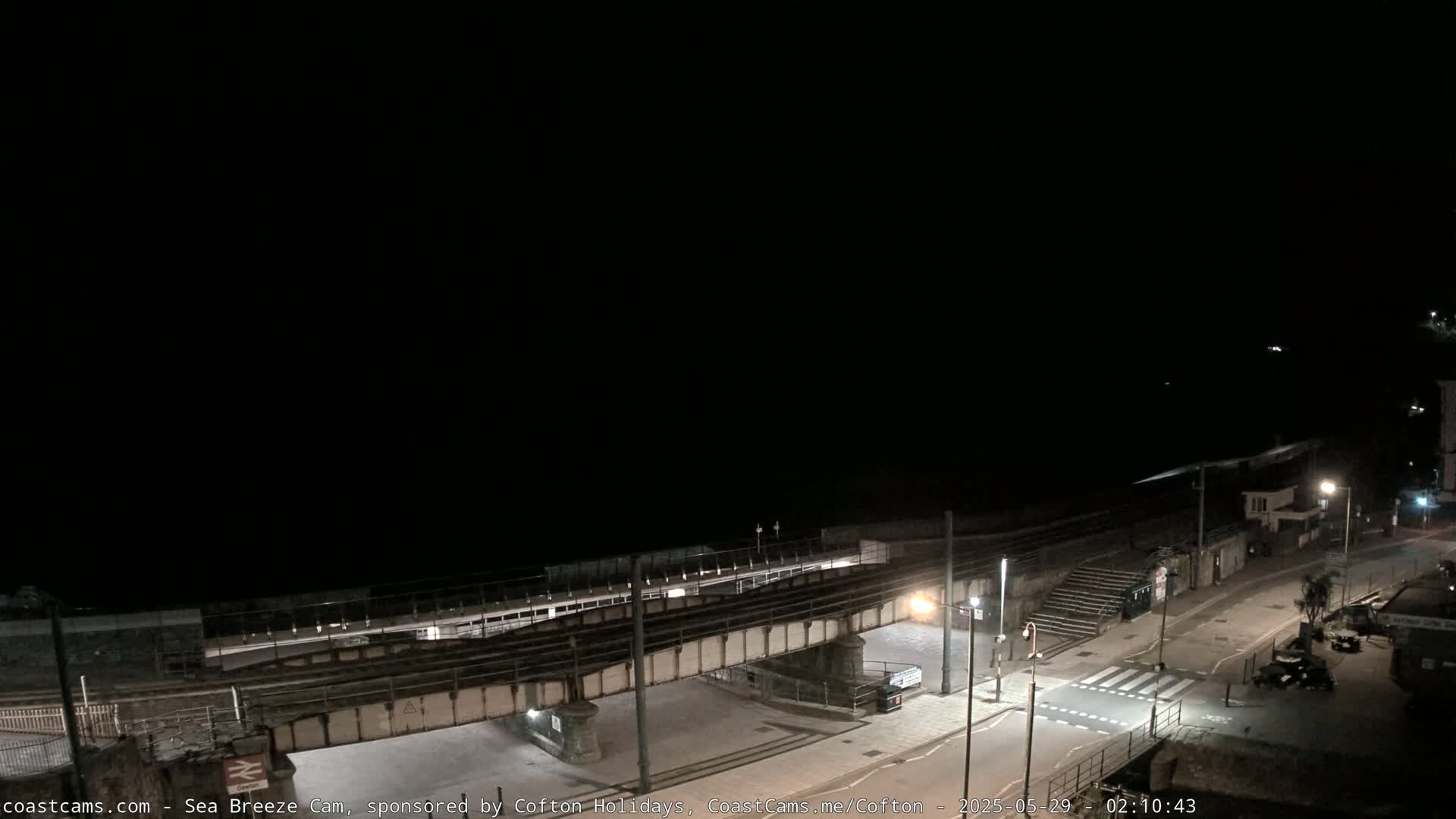 A nighttime, overhead view of a coastal train station and road, under a clear, dark sky.