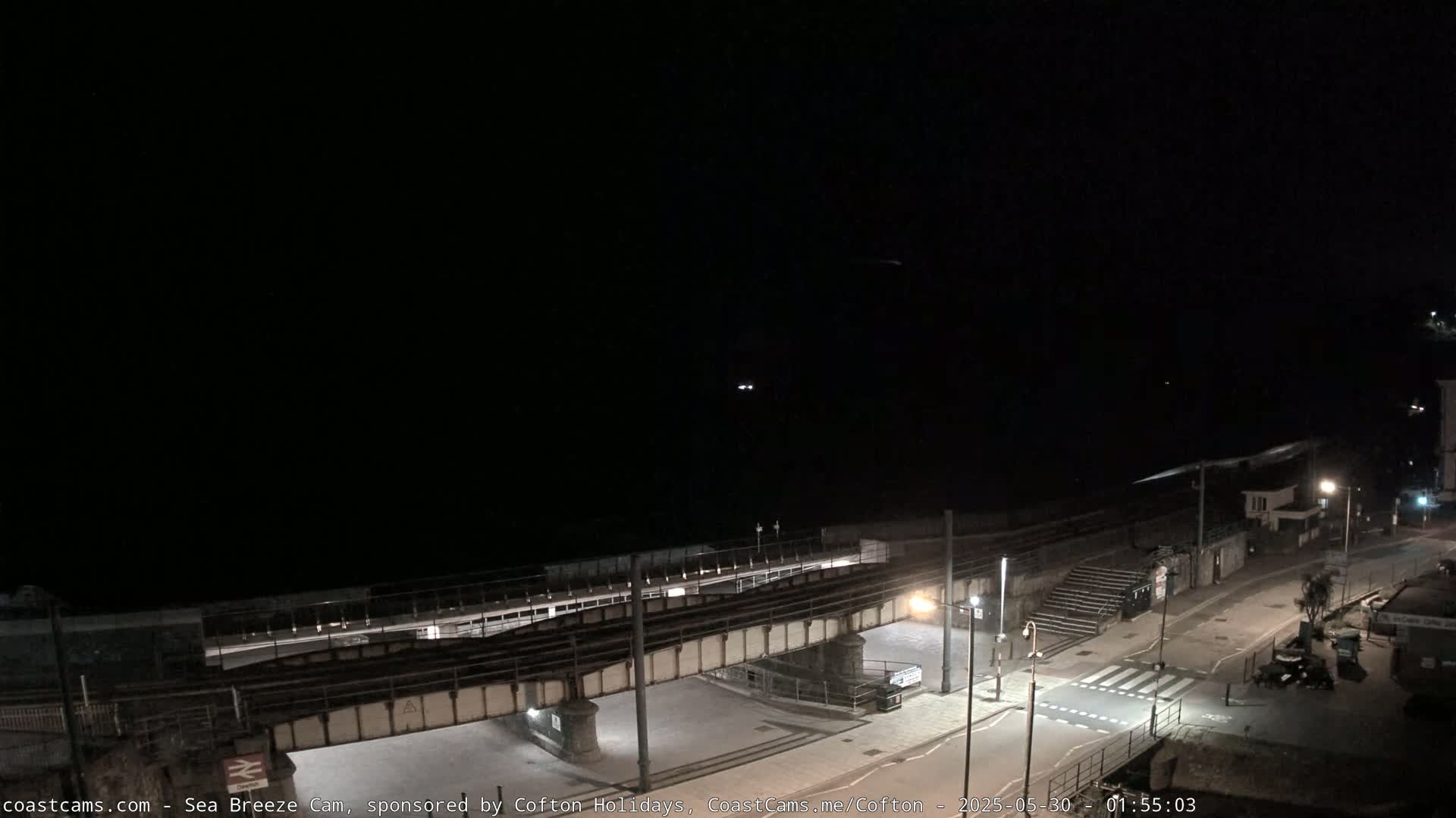 A nighttime view of a coastal town, showing a railway bridge over a road leading to a beach under a dark, clear sky.