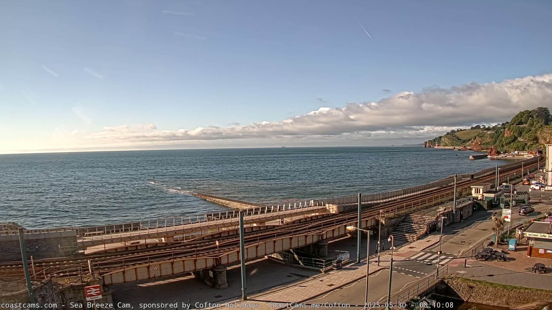 A coastal scene with partly cloudy skies shows a railway line running along a sea wall beside a beach and a town.