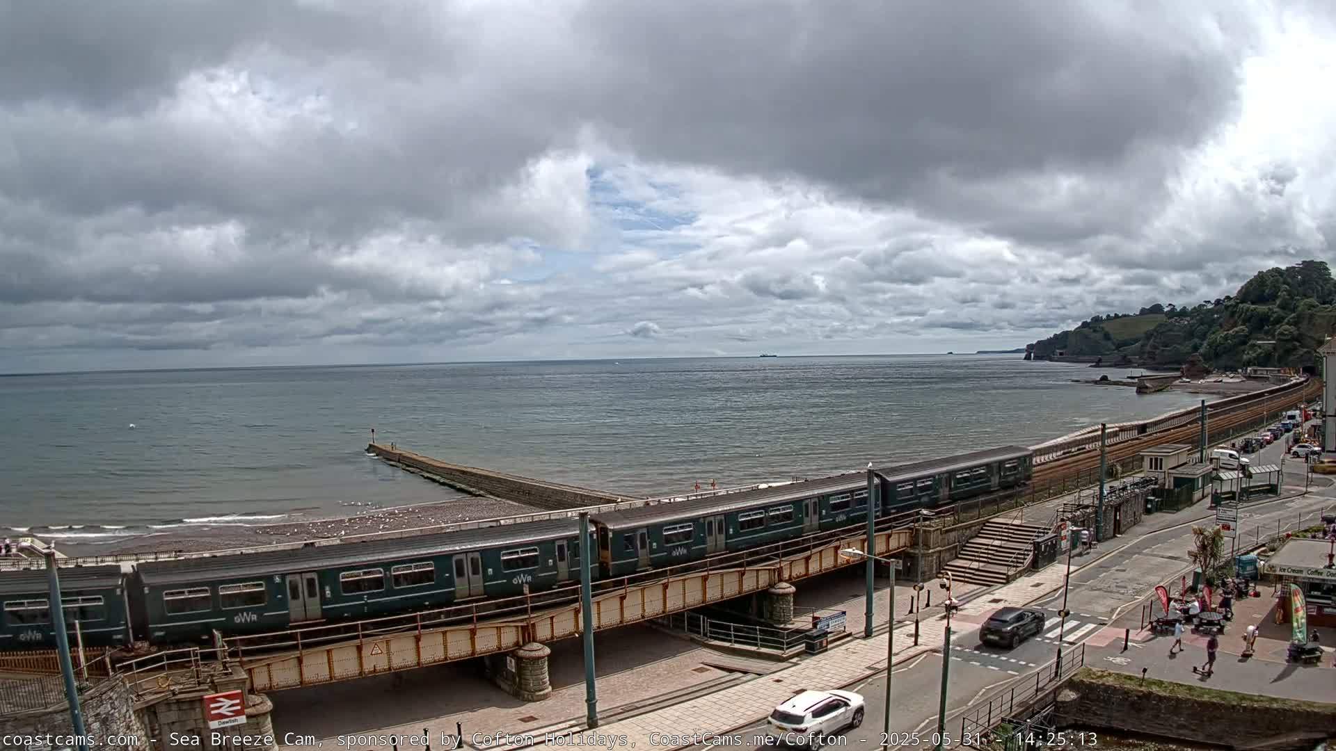 A green passenger train crosses a bridge over a coastal road, with a mostly cloudy sky and calm sea visible in the background.