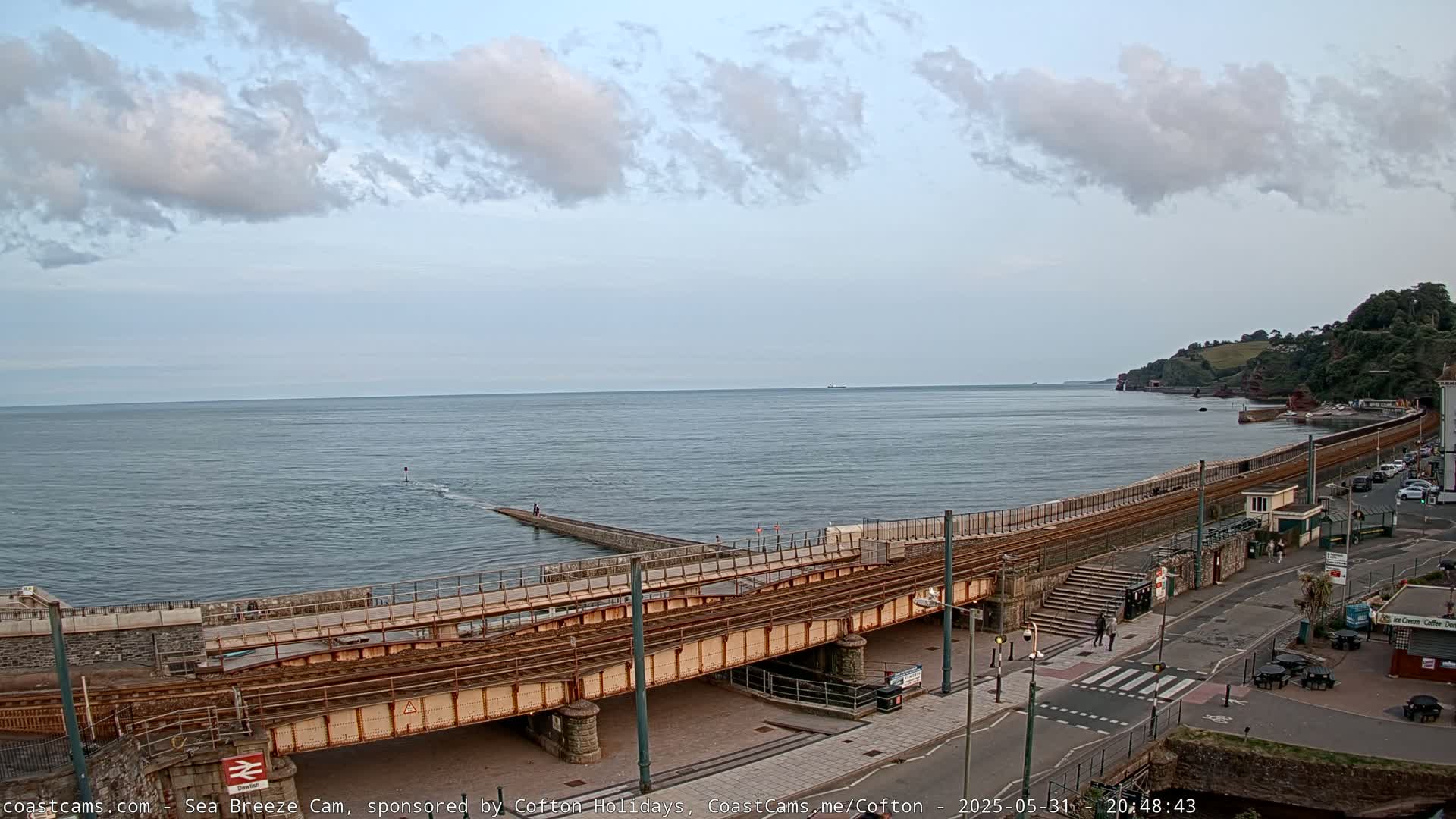 A railway line crosses a stone bridge over a coastal road, with a calm sea and partly cloudy sky in the background.
