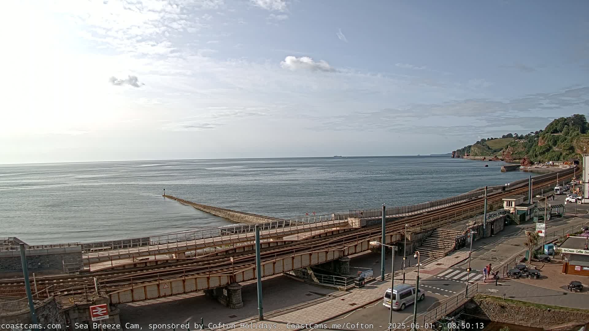 A coastal scene on a partly sunny day shows train tracks crossing a bridge over a seaside road, leading to a calm ocean with a distant headland.