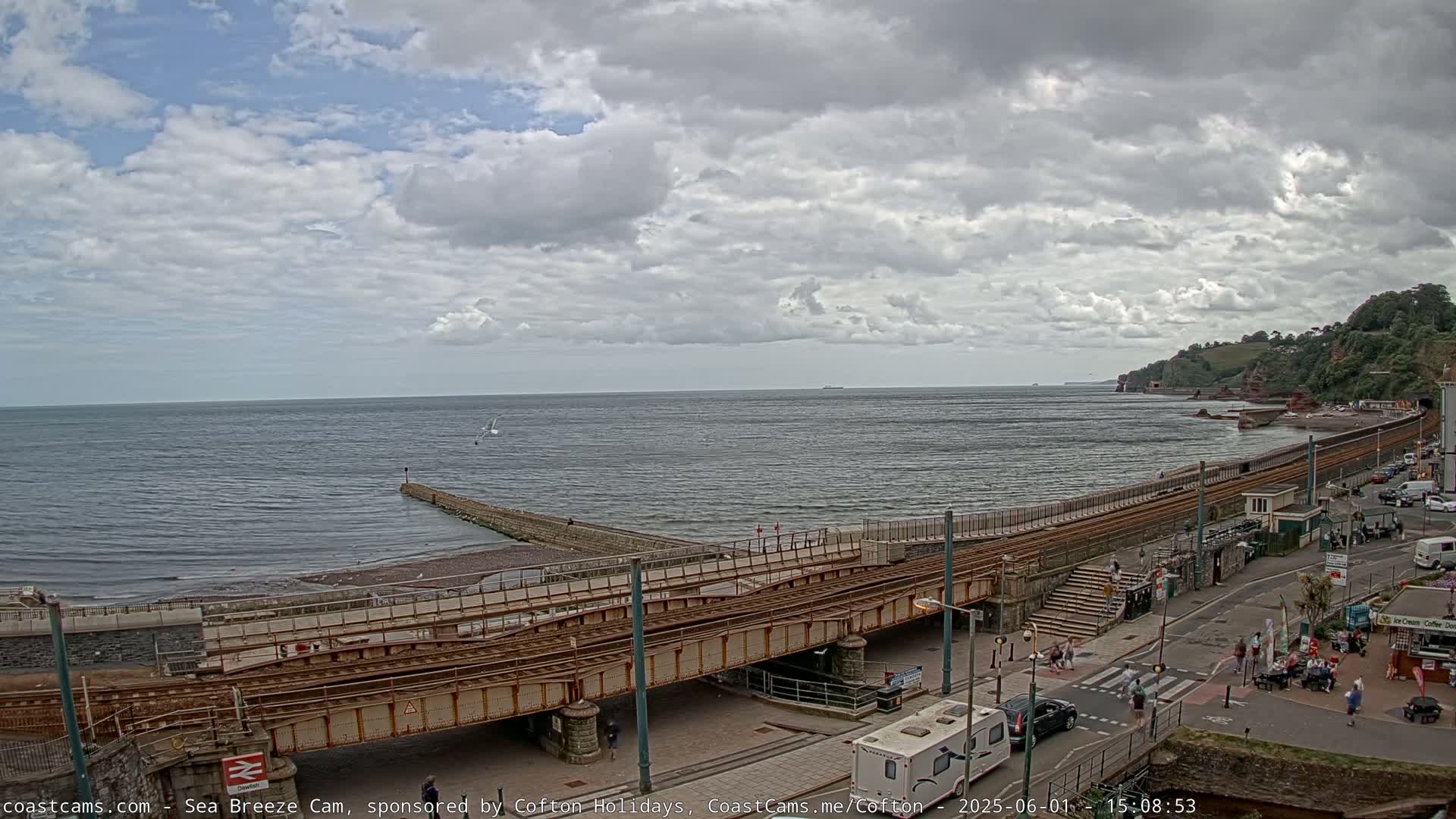 A coastal scene under a partly cloudy sky shows a railway track running alongside a beach and a road, with people and vehicles visible.