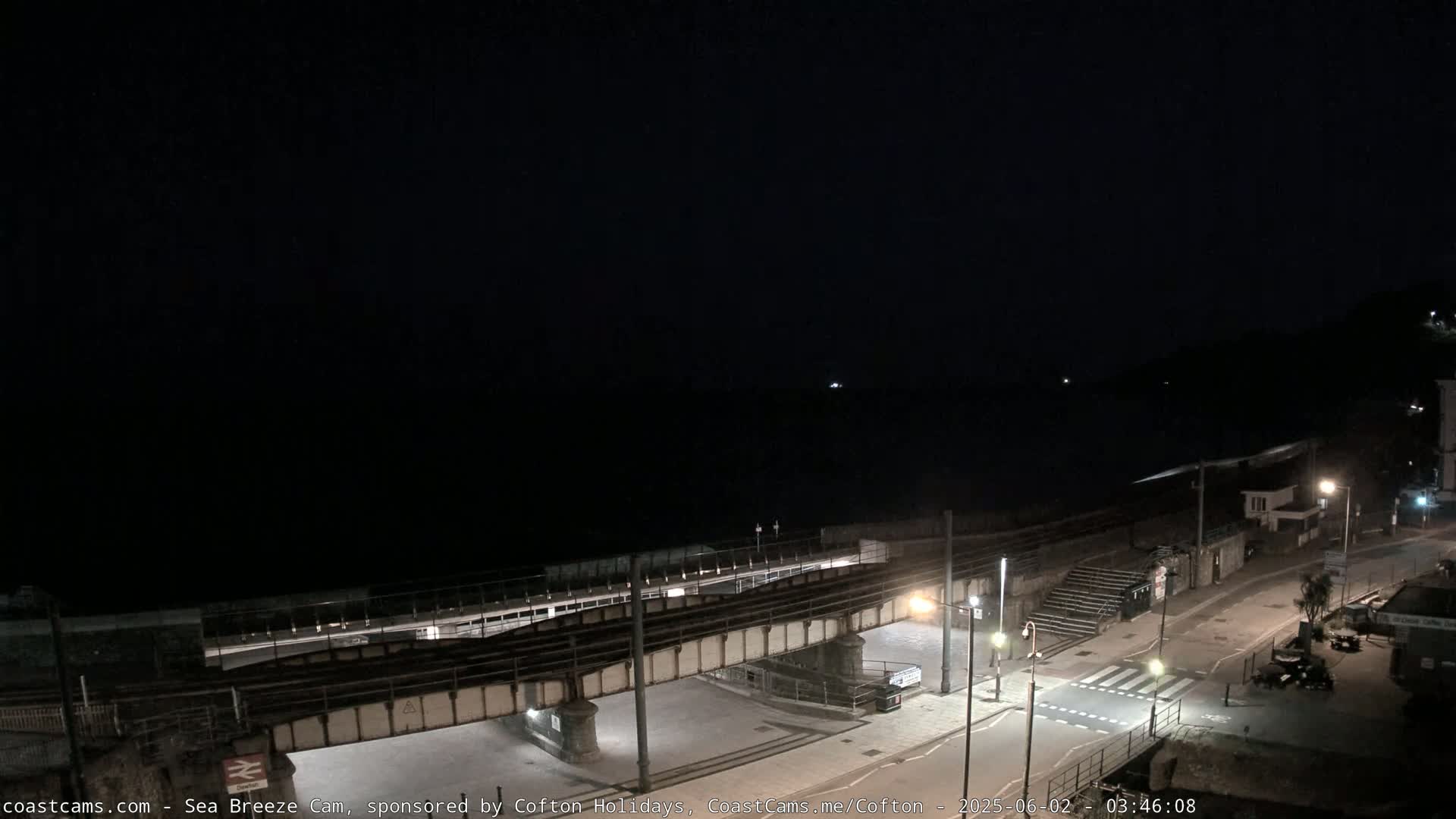 A nighttime view of a coastal town, showing a dark sea, a railway bridge over a roadway, and dimly lit streets.