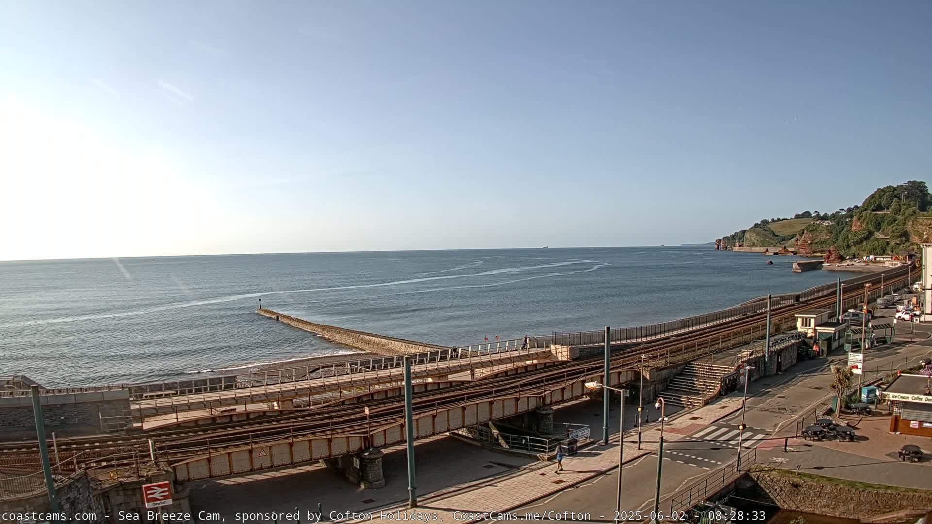A mostly sunny day reveals a coastal scene with train tracks crossing a stone embankment above a beach, alongside a calm ocean and reddish cliffs in the distance.