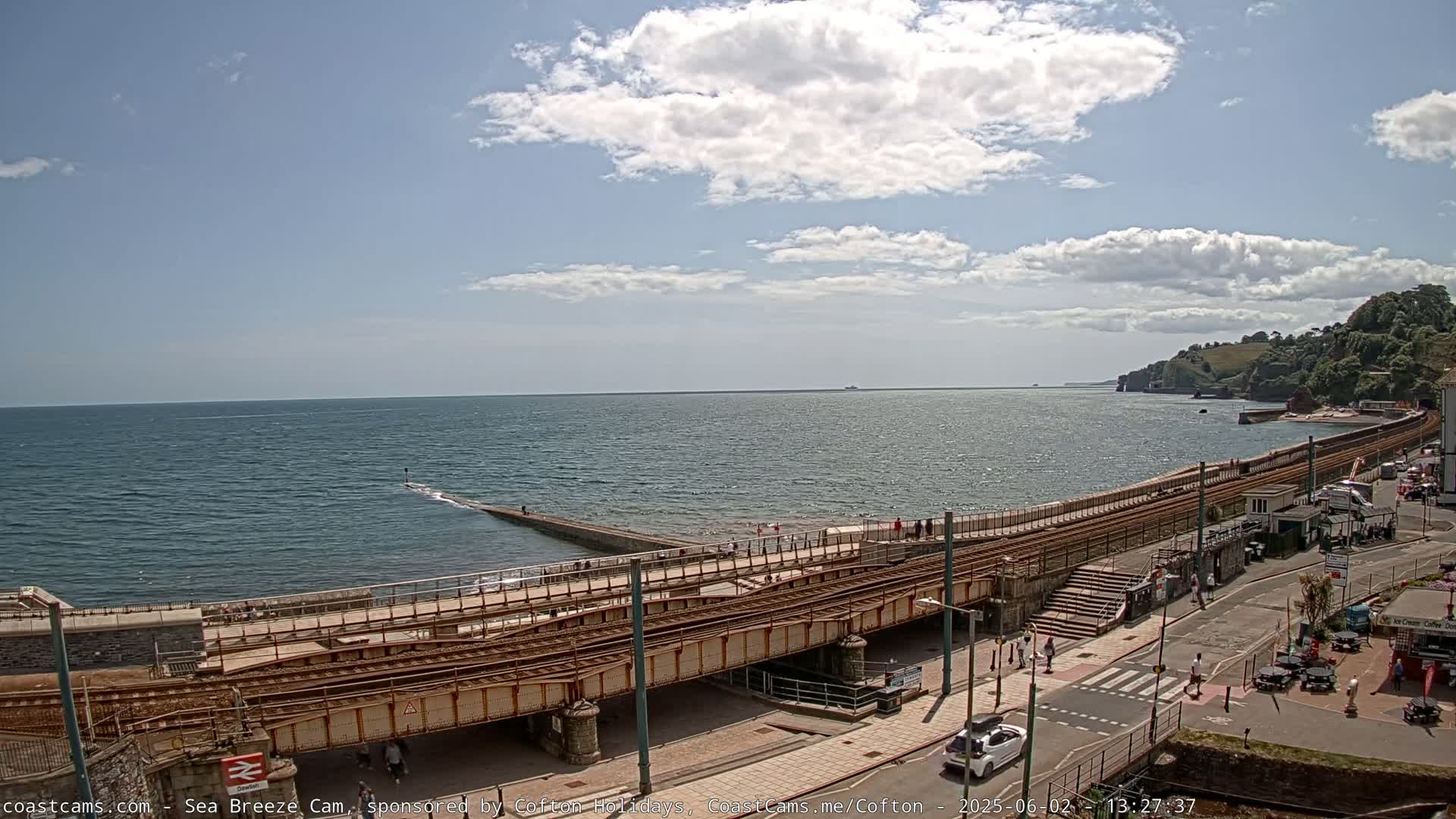 A mostly sunny day shows a coastal town with a train track running along a seawall, and people walking along the wall and streets.