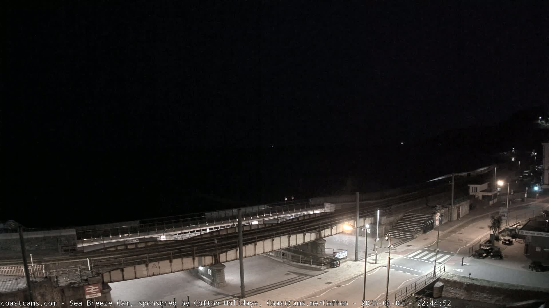 A nighttime view of a coastal town, showing a train trestle over a road and a mostly dark beach and sea.