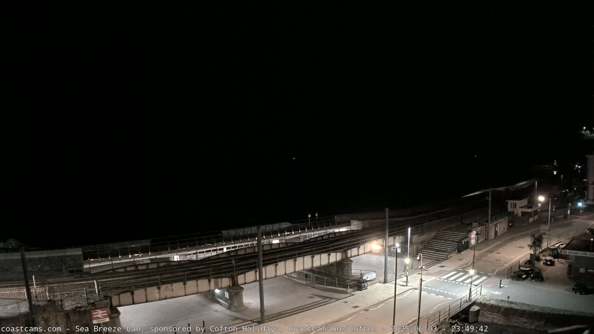 A nighttime coastal scene shows a train trestle over a road leading to the beach, with a few lights visible and a dark sky.