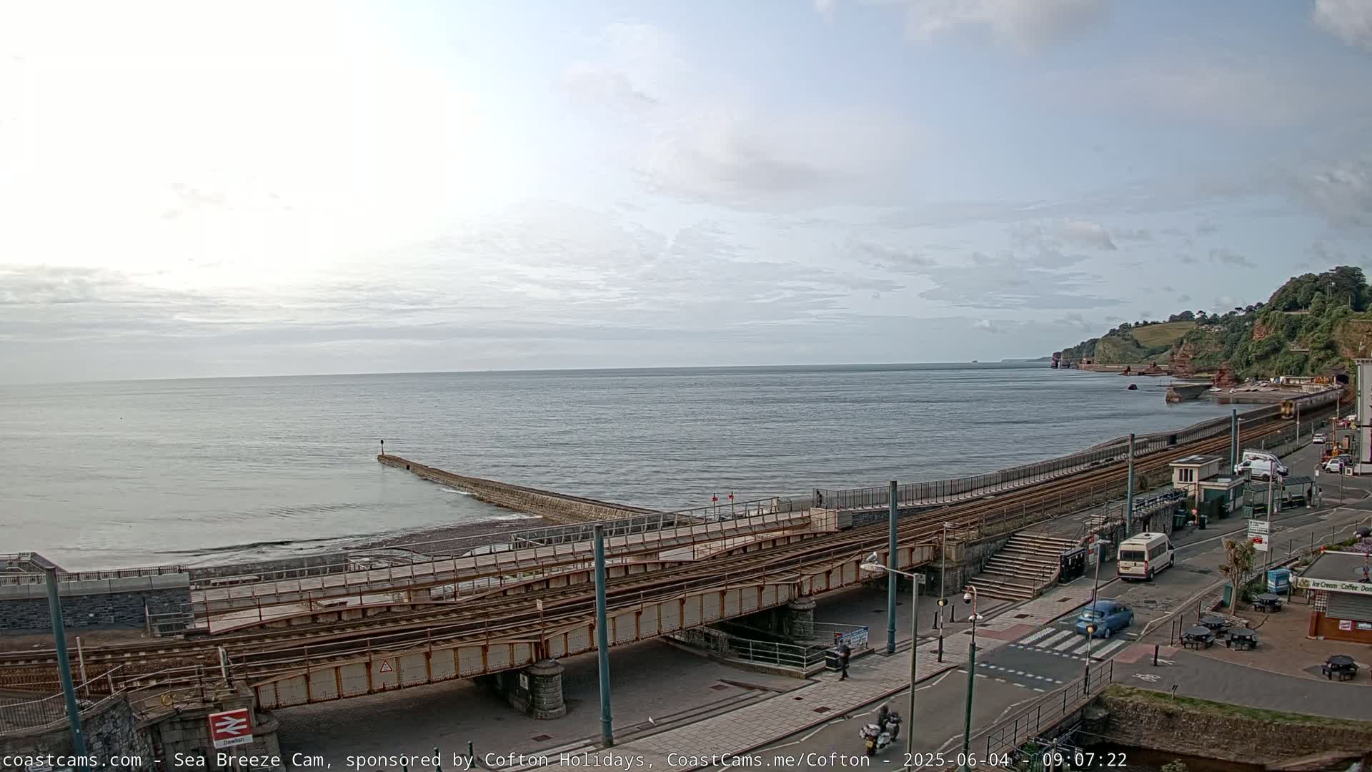 A mostly cloudy day overlooks a calm ocean with a railway crossing over a beach and road.