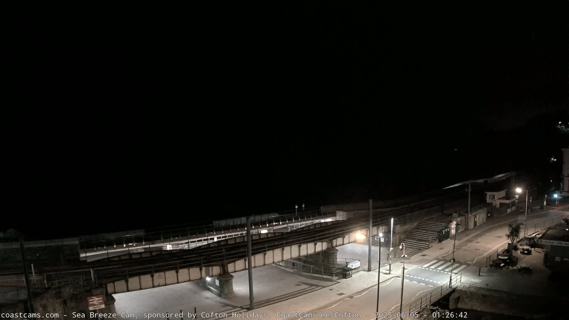 A dark nighttime scene shows a coastal road, a railway bridge, and a mostly empty beach under a clear, black sky.