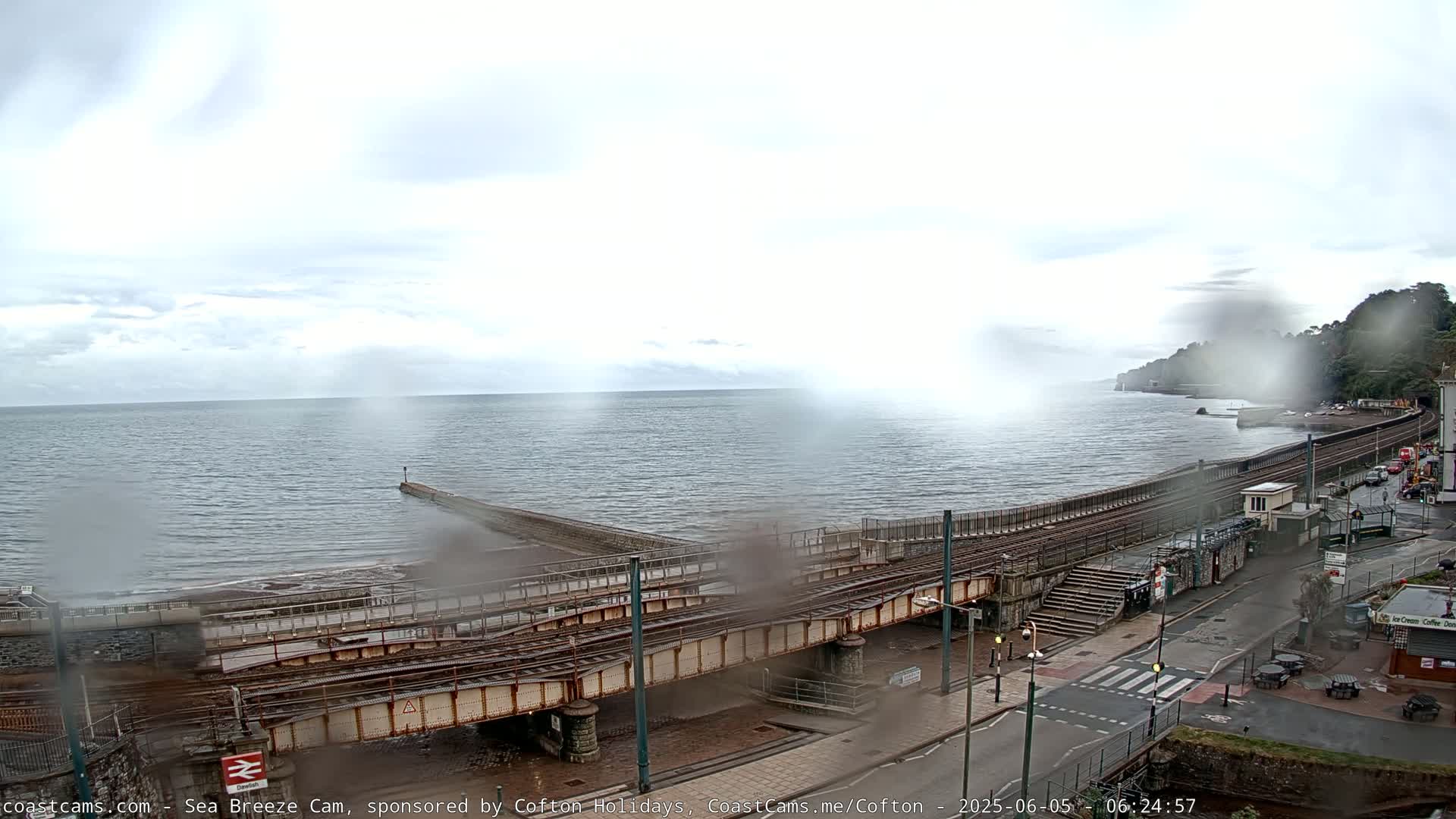 A rainy seaside town is viewed from above, showing train tracks crossing a road near the ocean, with a breakwater extending into the water.