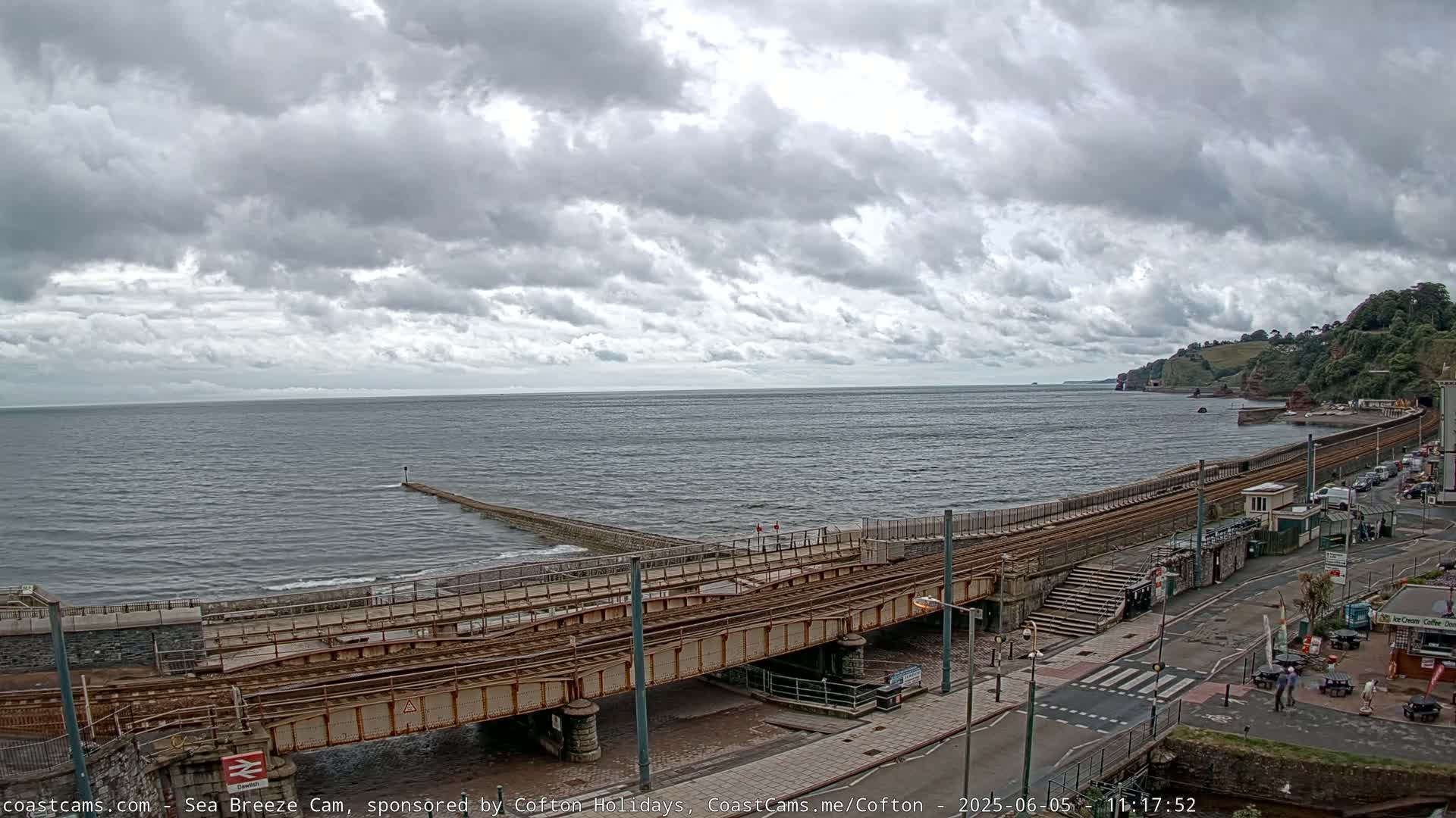 A coastal scene under a cloudy sky shows train tracks extending along a seawall next to a road, with a body of water and distant cliffs in the background.