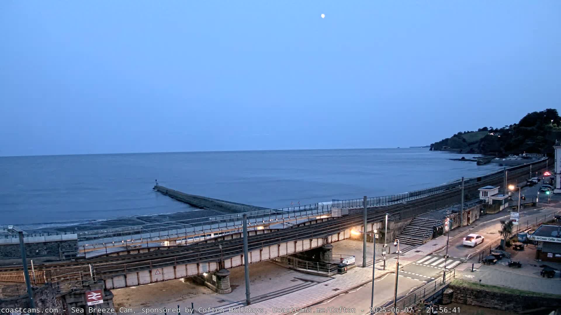 A train track crosses over a coastal road near a calm sea under a twilight sky with a visible moon.