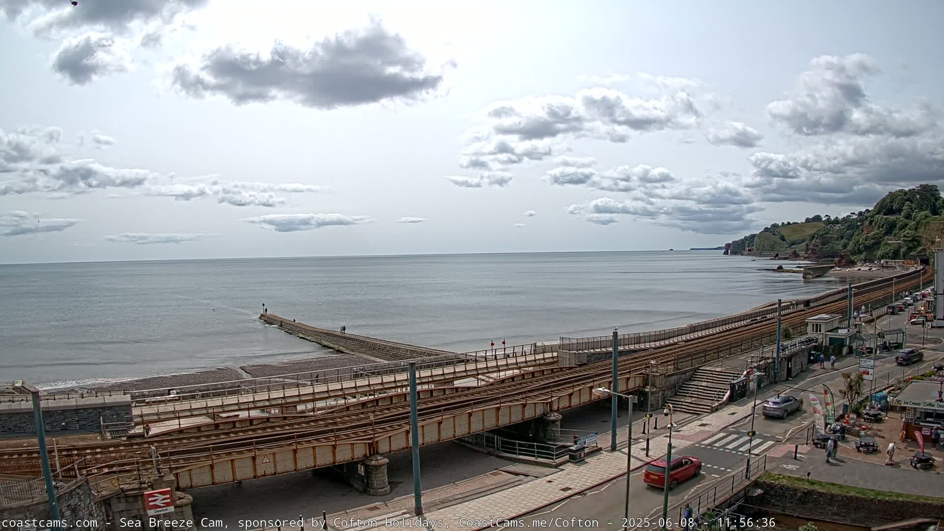 A coastal scene on a partly cloudy day shows a beach with a railway line crossing it, leading to a town with buildings and cars along the shoreline, with cliffs visible in the distance.