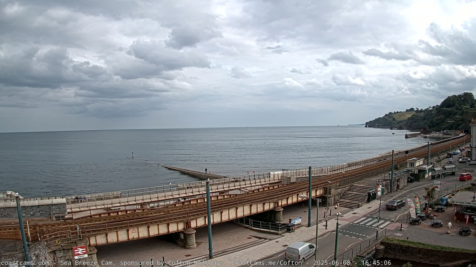 A coastal scene under a cloudy sky shows a railway line running along a seawall, with a town visible beyond.