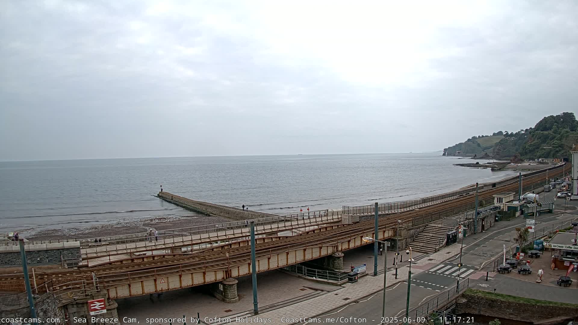 A coastal scene under an overcast sky shows a railway line running along a beach next to a calm sea with a breakwater and a small town visible in the background.