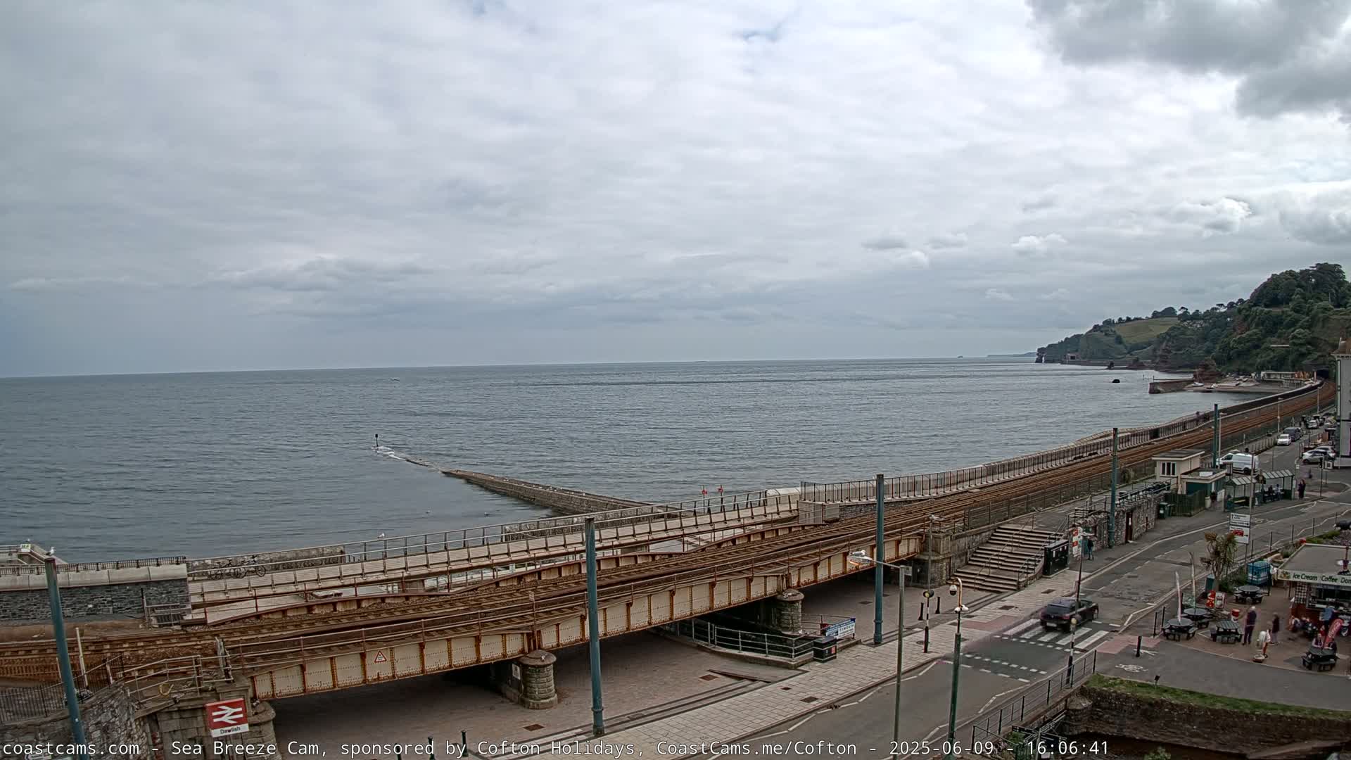 A coastal scene under an overcast sky shows a railway line and bridge crossing over a road next to the sea, with a small town visible in the distance.
