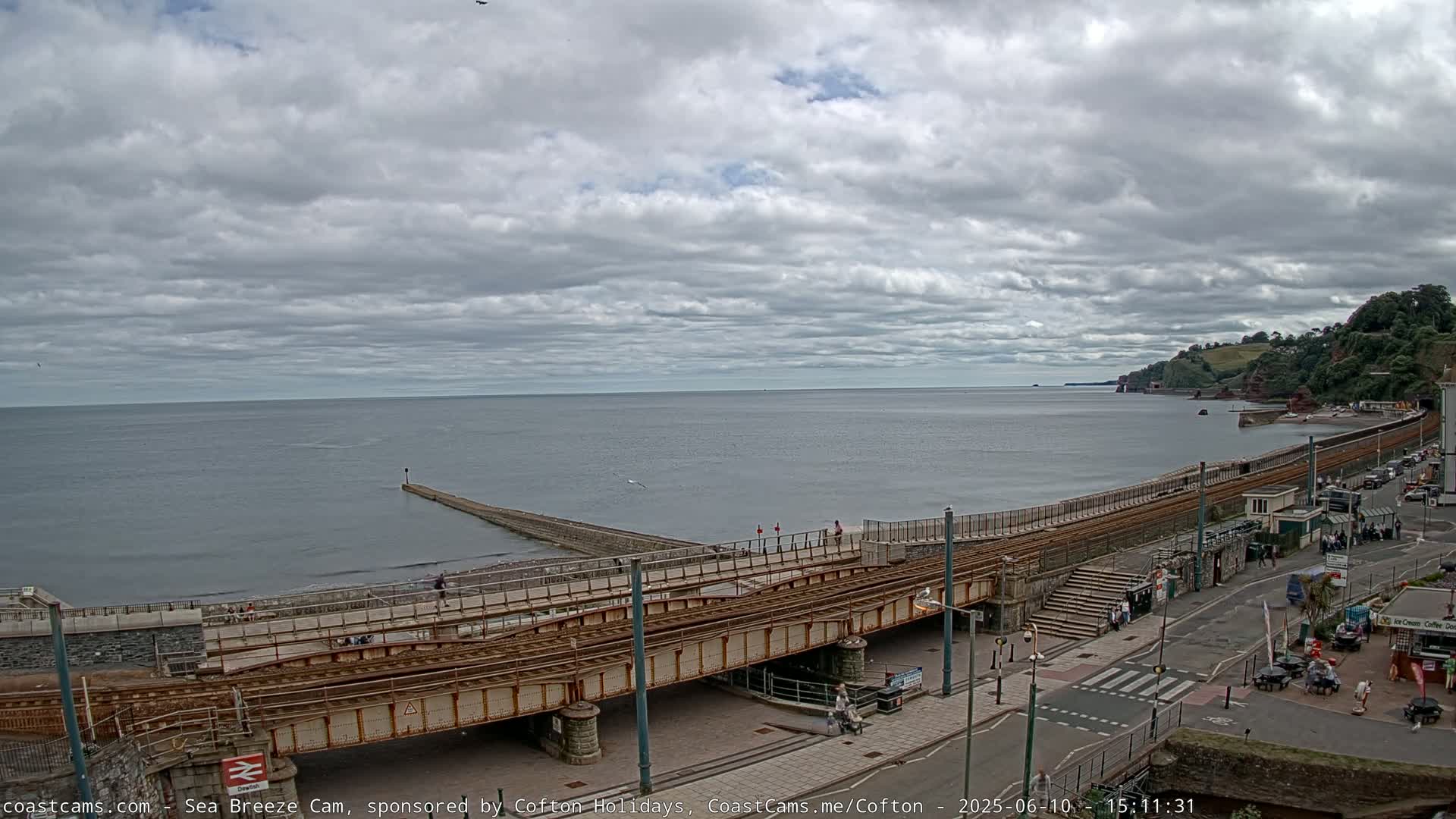 A coastal scene under a cloudy sky shows a railway track running alongside a beach and curving towards a headland, with a pier extending into the calm sea.