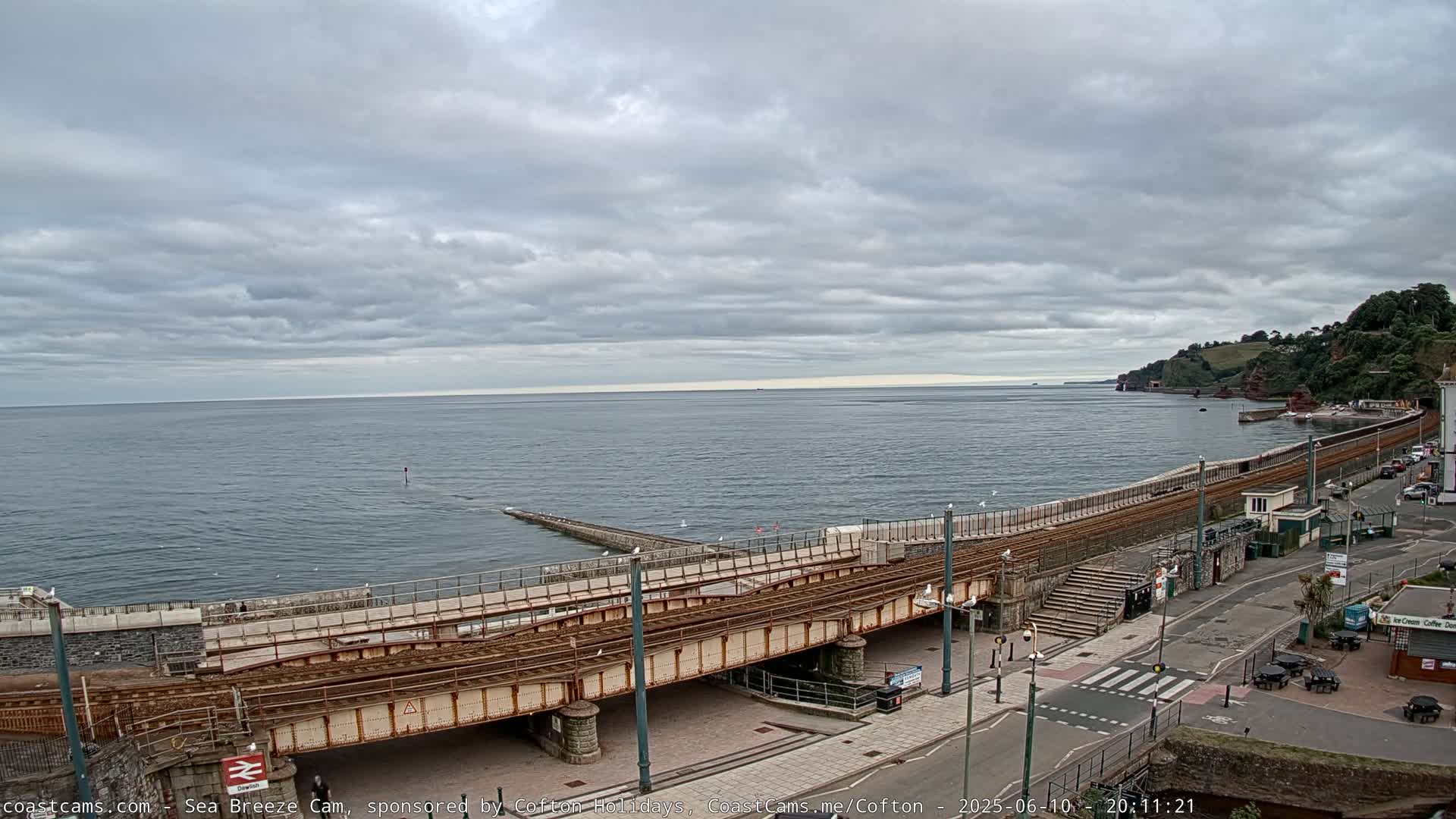 A coastal scene under a cloudy sky shows train tracks curving along a seawall next to a road with buildings and parked cars.