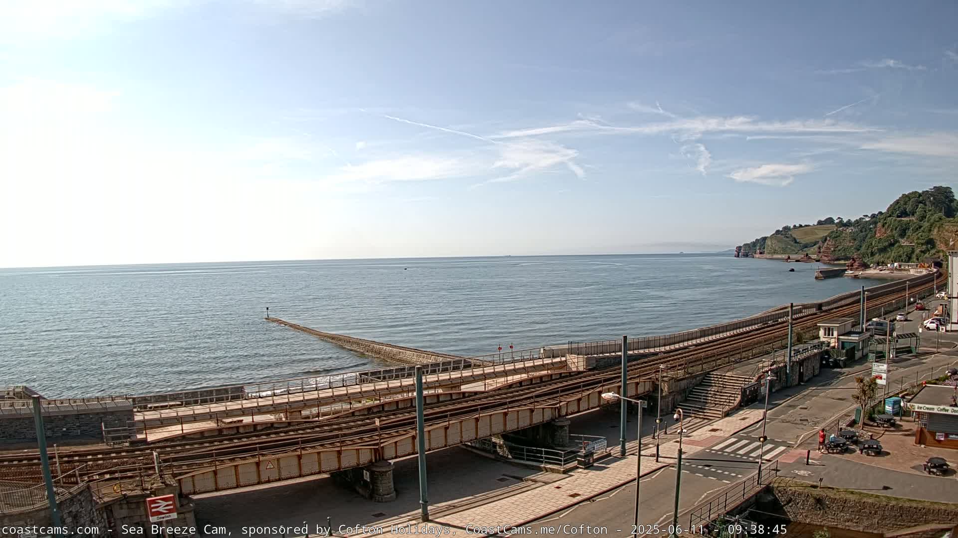 A coastal railway line and adjacent road run alongside a calm sea under a mostly clear sky.