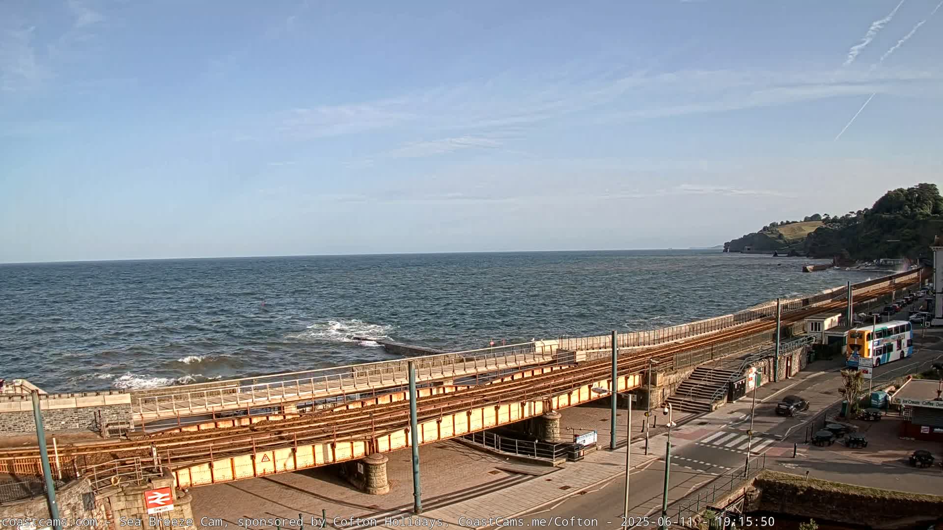 A seaside town scene with a slightly choppy sea under a mostly clear sky shows train tracks running alongside a coastal road, with a bus and cars visible.