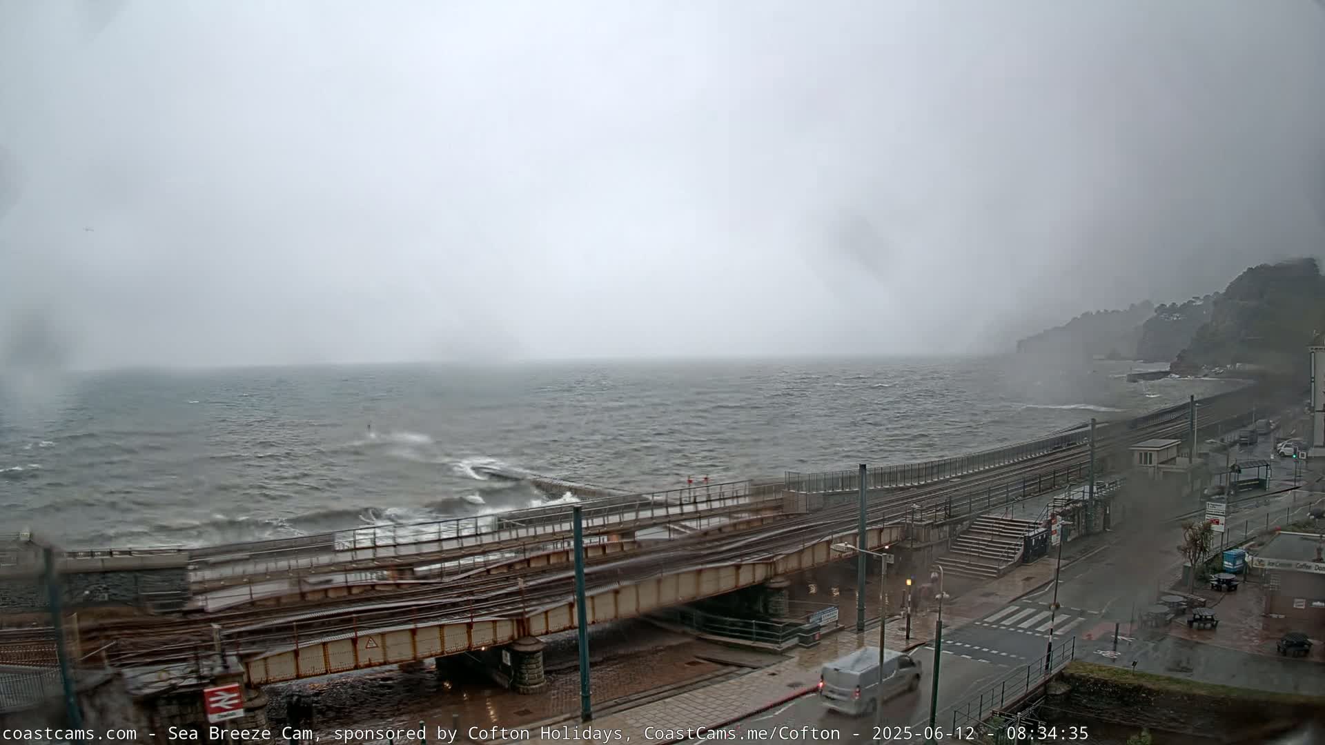 A stormy, overcast day shows a choppy sea crashing against a seawall near a train trestle and coastal road.