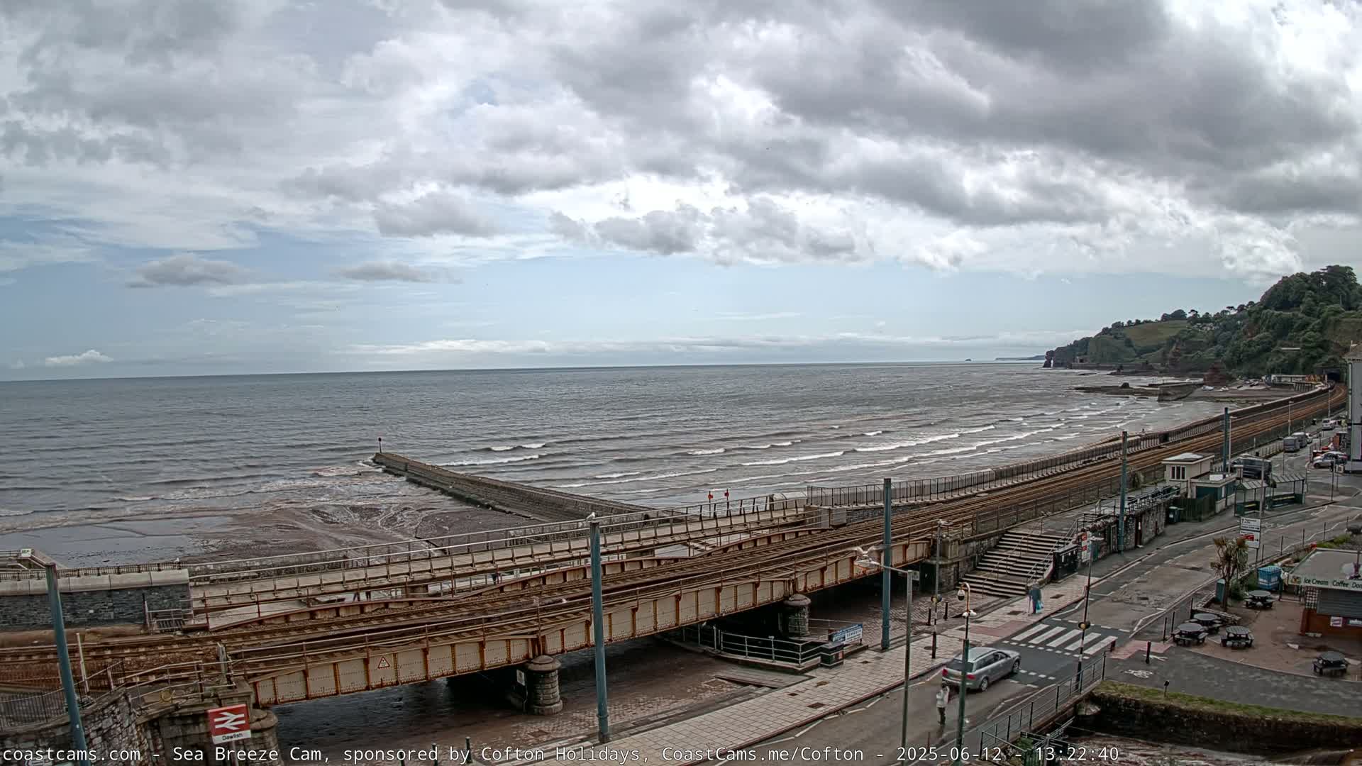 A partly cloudy day shows a beach with moderate waves, a railway line running along the shore, and a town beyond.