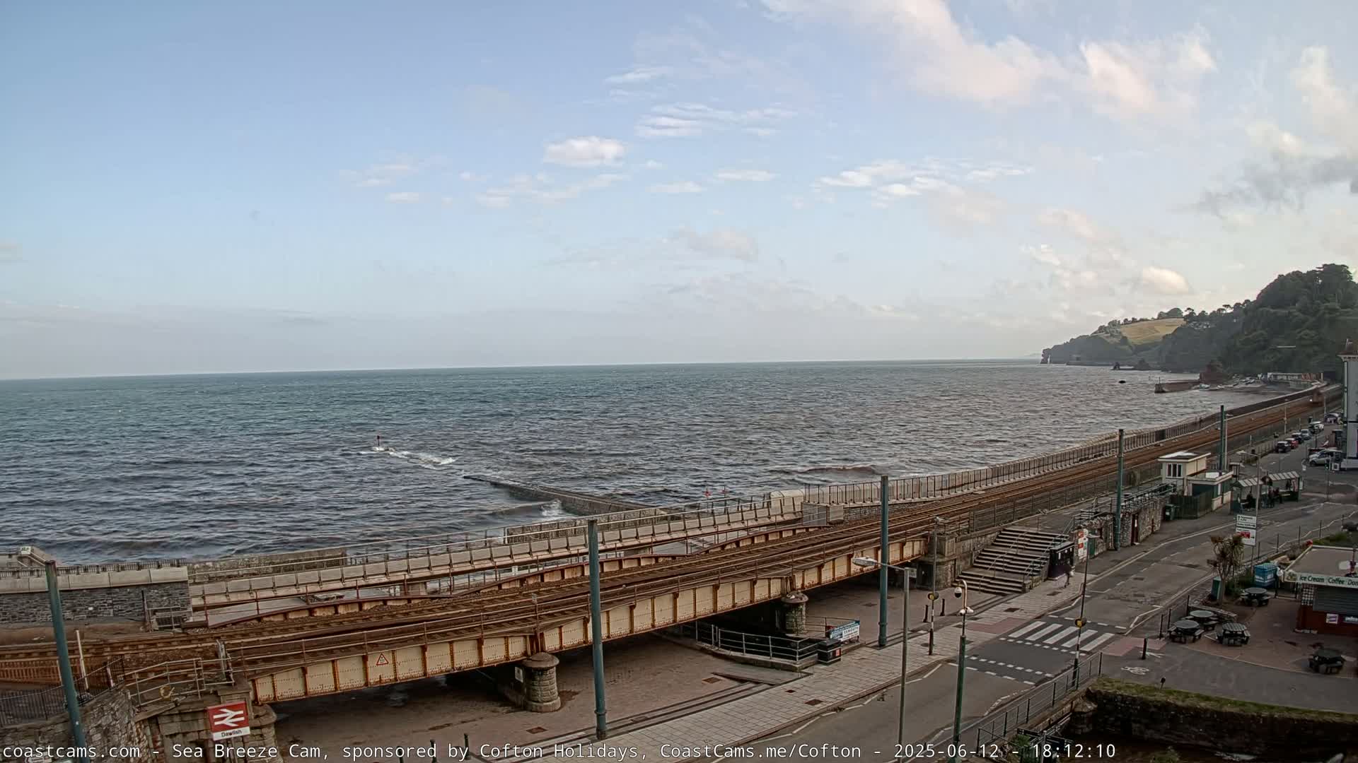 A slightly choppy sea stretches to a distant coastline under a partly cloudy sky, with a railway line running along a seawall next to a road.