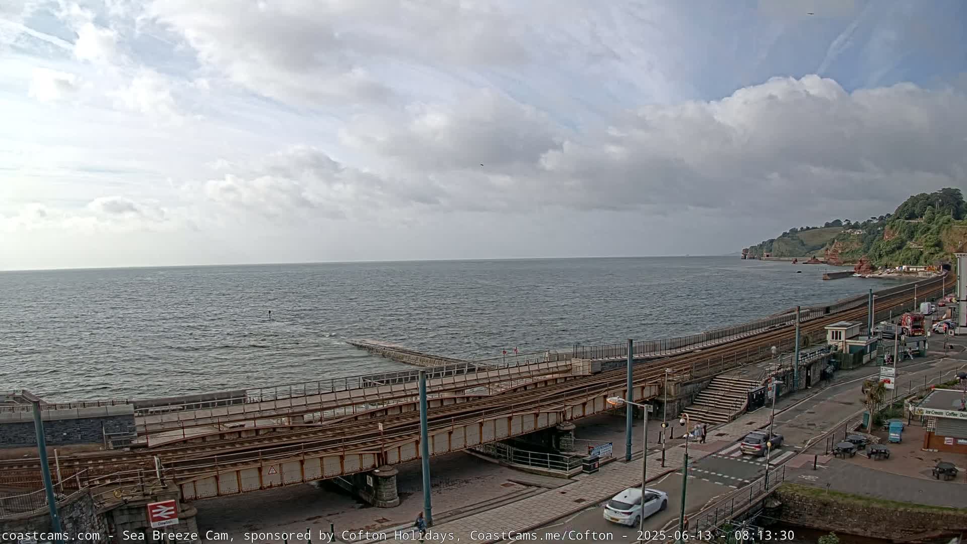 A partly cloudy day overlooks a choppy sea with a railway track and coastal road leading to a cliffside.