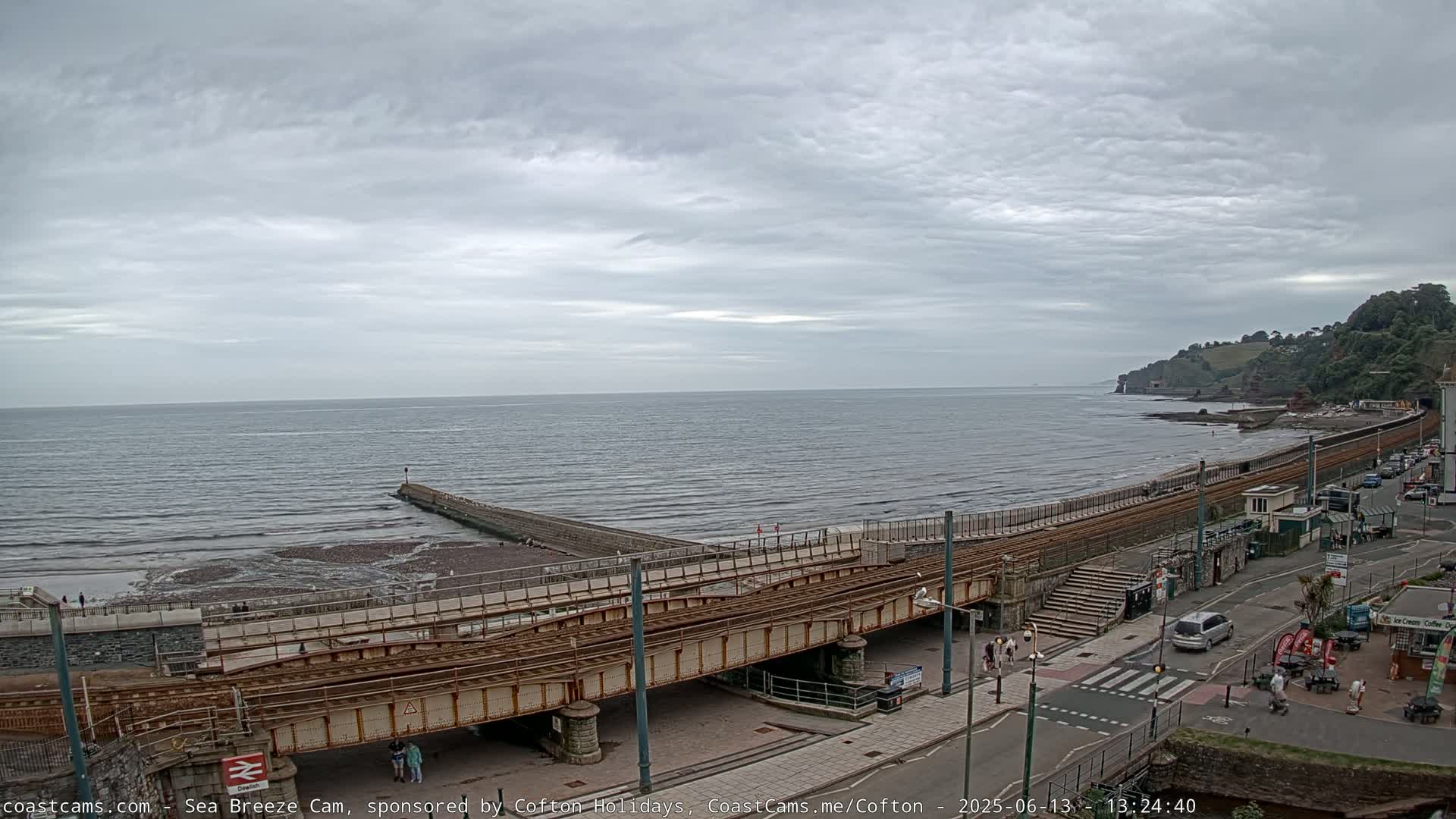 A coastal scene under an overcast sky shows a railway track crossing a road above a beach, with a calm sea stretching to the horizon and a small pier jutting out into the water.