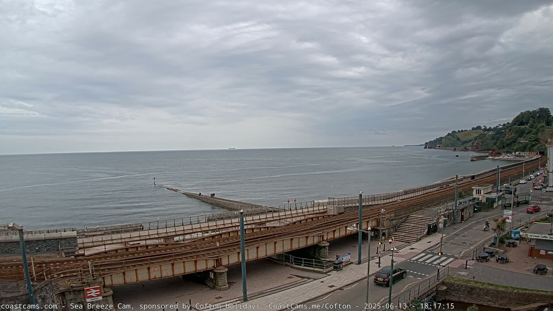 A coastal scene under an overcast sky shows a railway line running along a seawall, with a calm sea and distant hills visible.