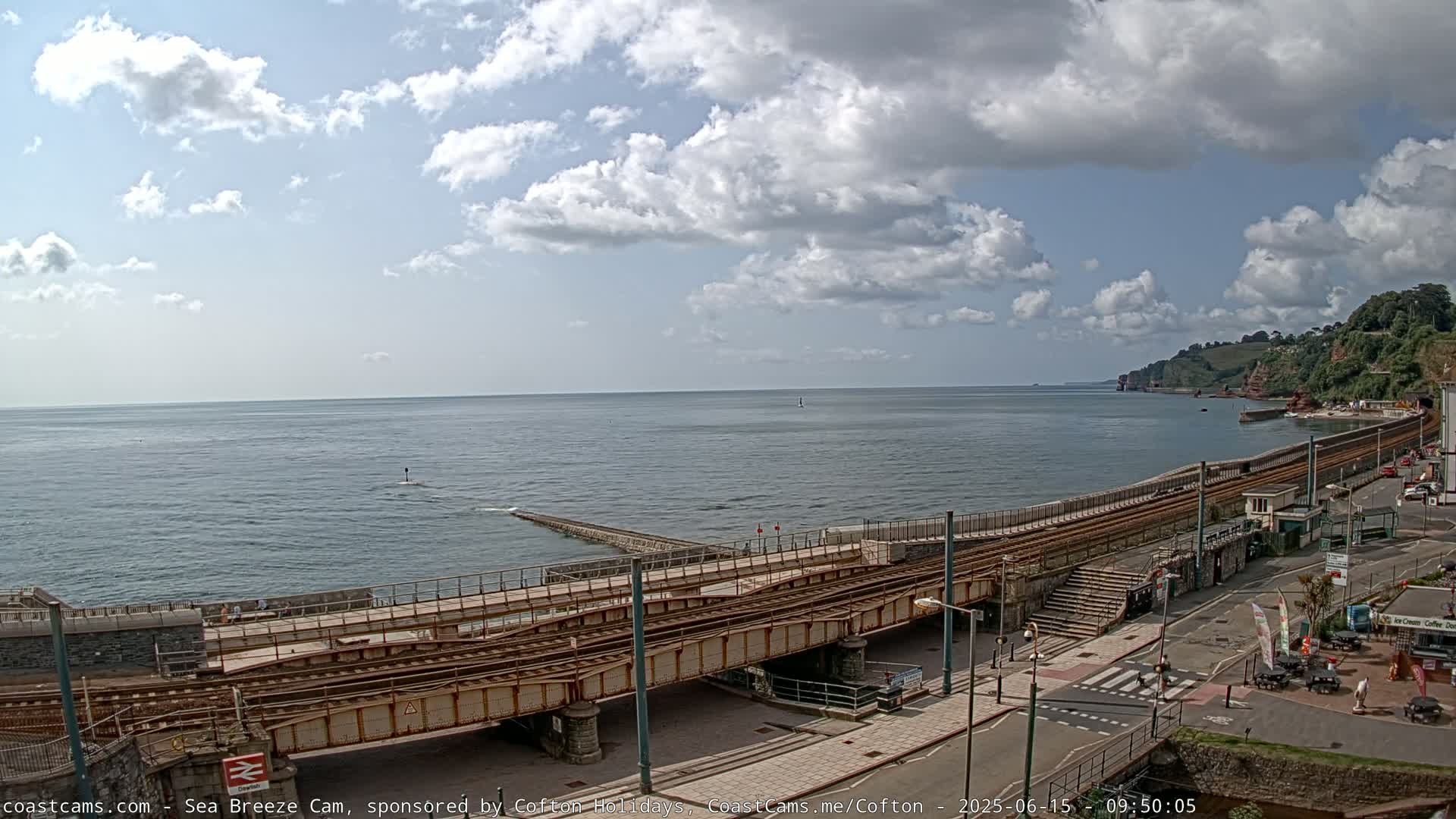 A partly cloudy day reveals a coastal scene with train tracks running along a seawall, next to a road and buildings.