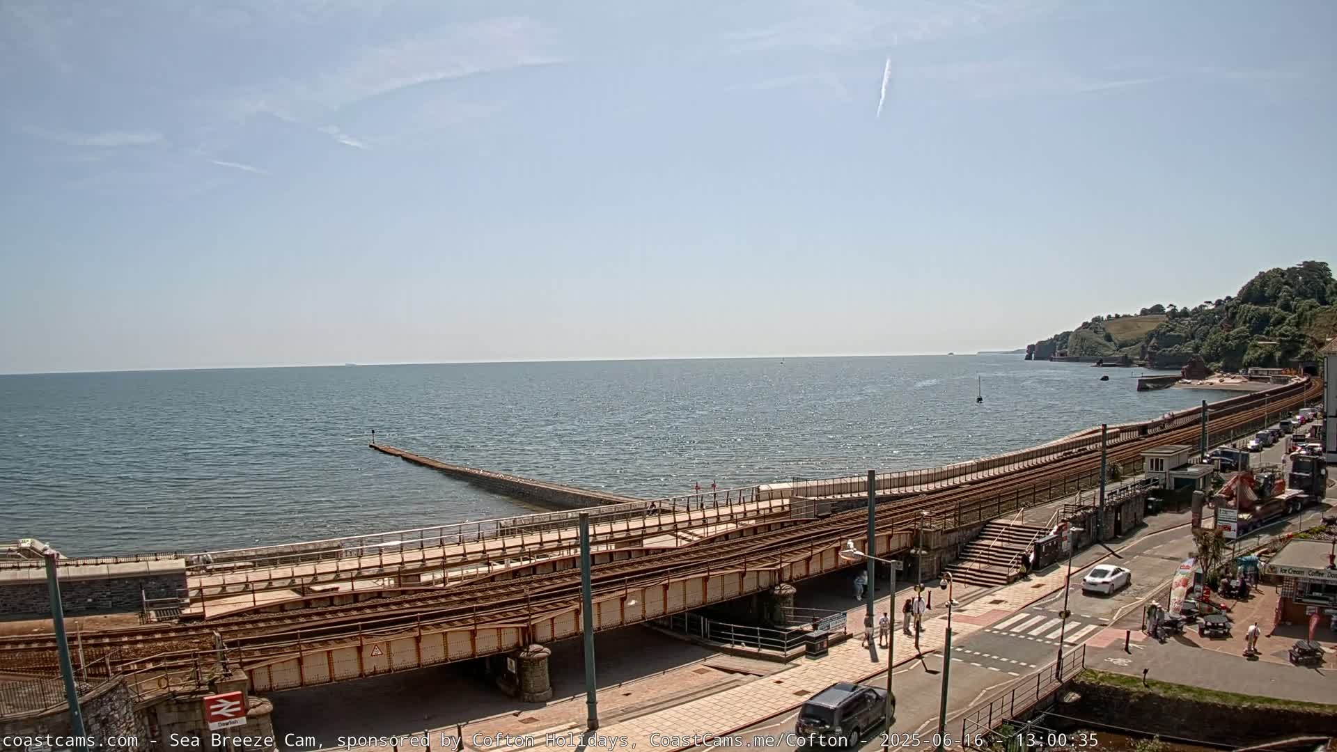 A sunny day reveals a coastal scene with a railway line traversing a seawall, alongside a road with cars and pedestrians, and a calm sea extending to a distant shoreline.